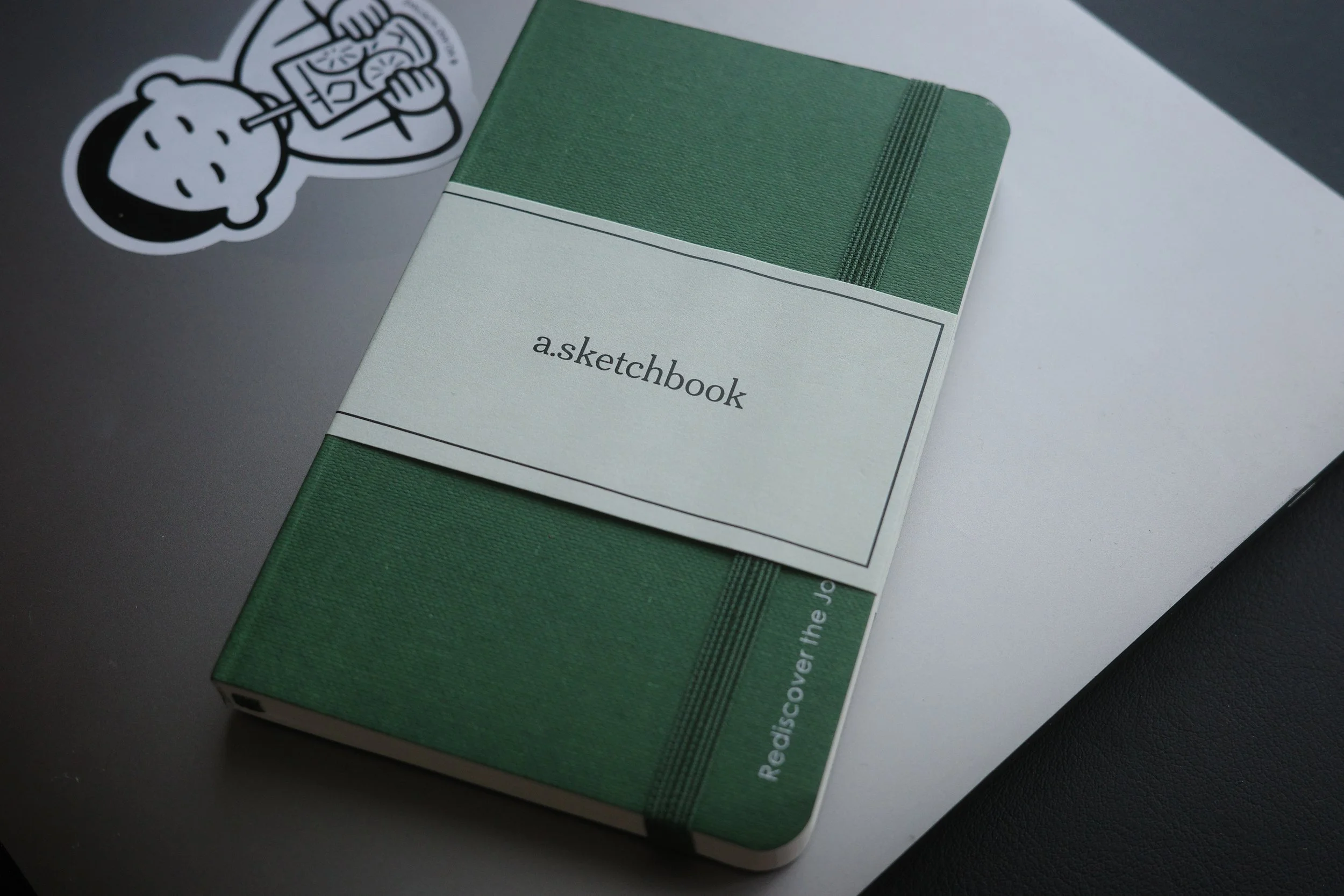 A green and white hardcover sketchbook titled 'a.sketchbook' resting on a silver MacBook laptop with a sticker of a cartoon bear holding a drink on it, on a white surface.