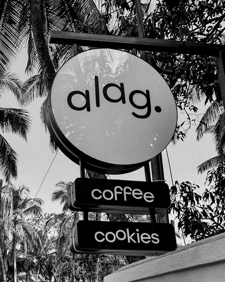 Sign for a coffee shop named alag. with additional signs below advertising coffee and cookies, set against a background of palm trees and sky.