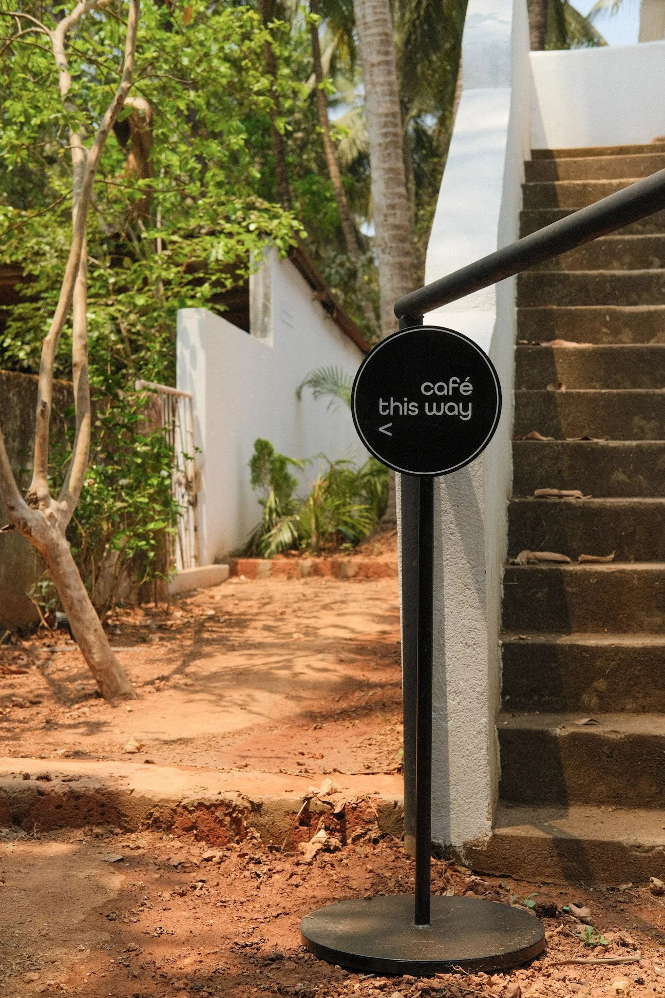 A black circular sign with white text indicating the direction to a café, placed near a white wall and brown stairs outdoors, with greenery and trees in the background.