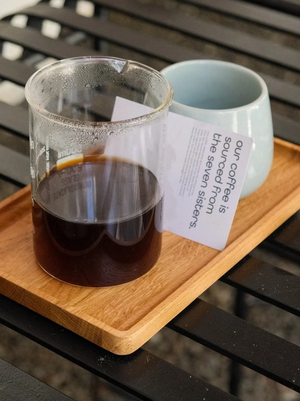 A glass of black coffee on a wooden tray next to a light blue mug and a note card that reads 'Our coffee is sourced from the seven sisters.' on a black outdoor table.