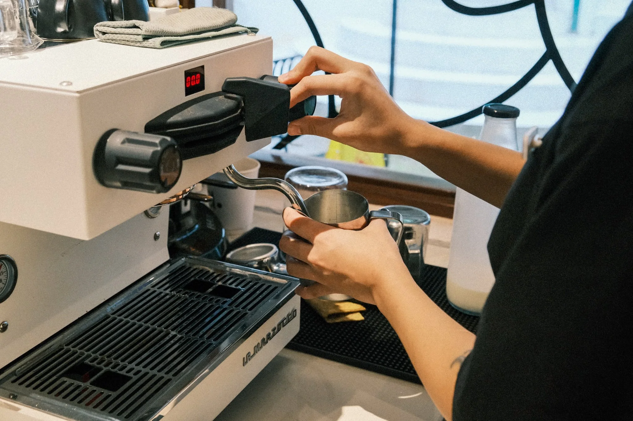 A person operating an espresso machine in a coffee shop, holding a metal pitcher under the espresso machine's steam wand.