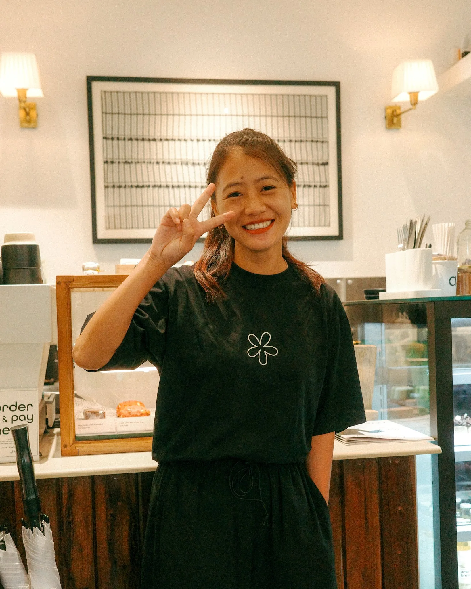 Smiling young woman with brown hair in a ponytail making a peace sign gesture inside a cafe.