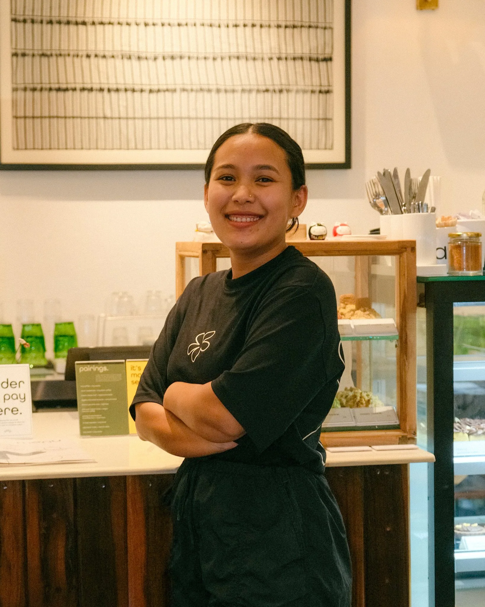 A smiling young woman with dark hair tied back, wearing a black T-shirt and black pants, standing with arms crossed inside a cafe or bakery.
