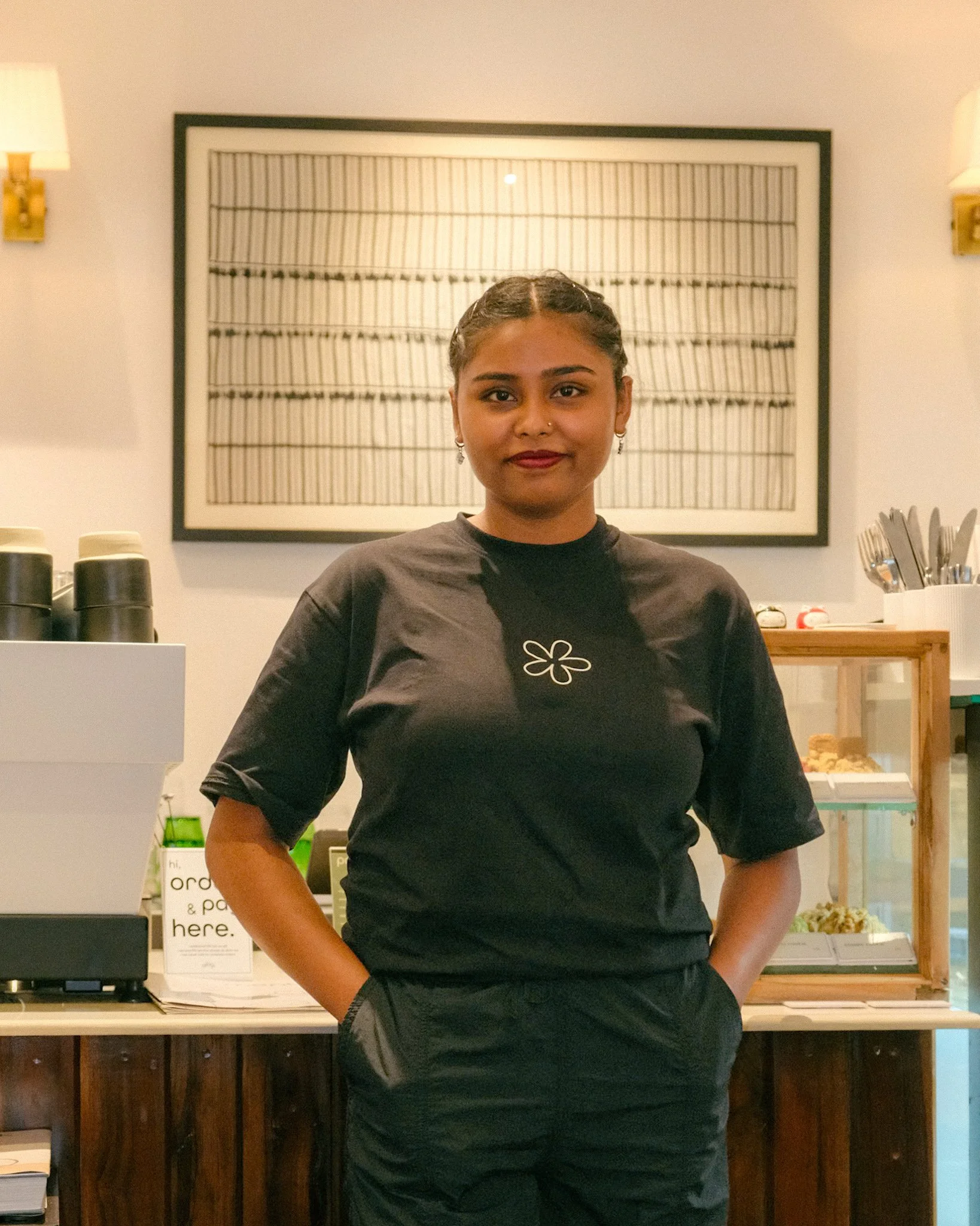 A young woman with dark hair in braids, wearing a black shirt and pants, standing inside a cafe with a framed artwork on the wall behind her, and a counter with cups, utensils, and food items.