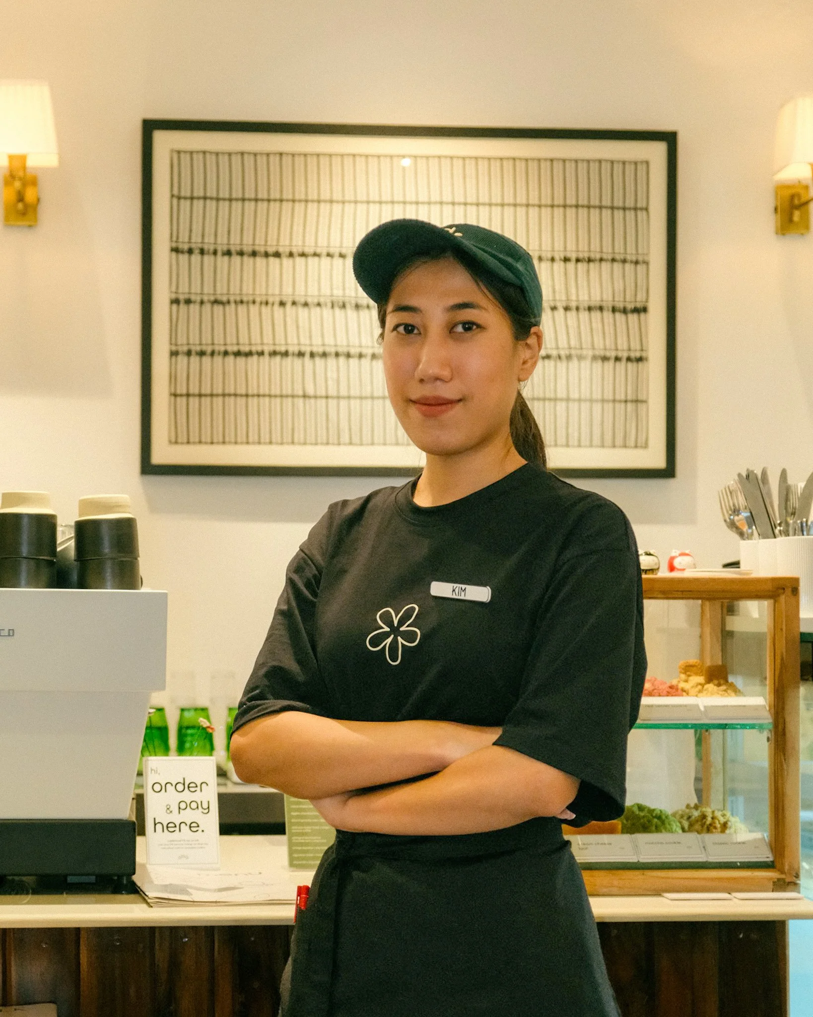 A young woman with a black cap and uniform standing behind a counter at a café, with arms crossed, smiling slightly.