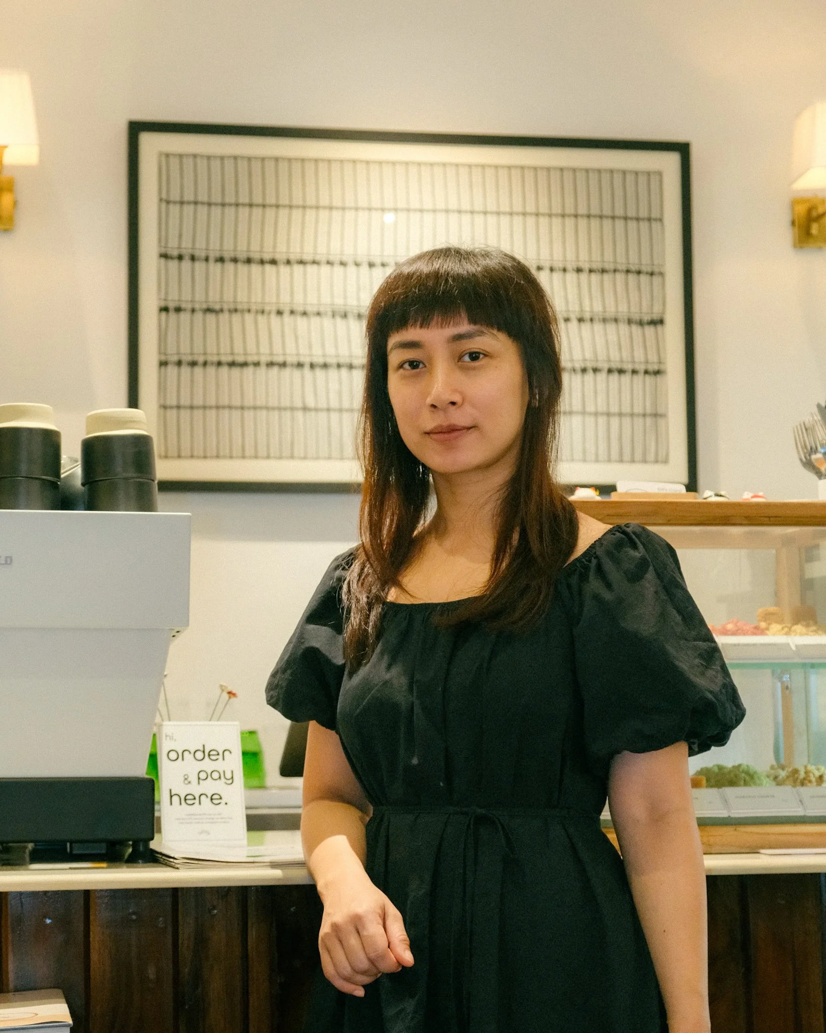 A woman standing inside a cafe or bakery, wearing a black dress with puffed sleeves, looking at the camera. Behind her is a white sign that says 'order & pay here,' and a display case with baked goods. The decor includes a large framed artwork and wall-mounted lamps.