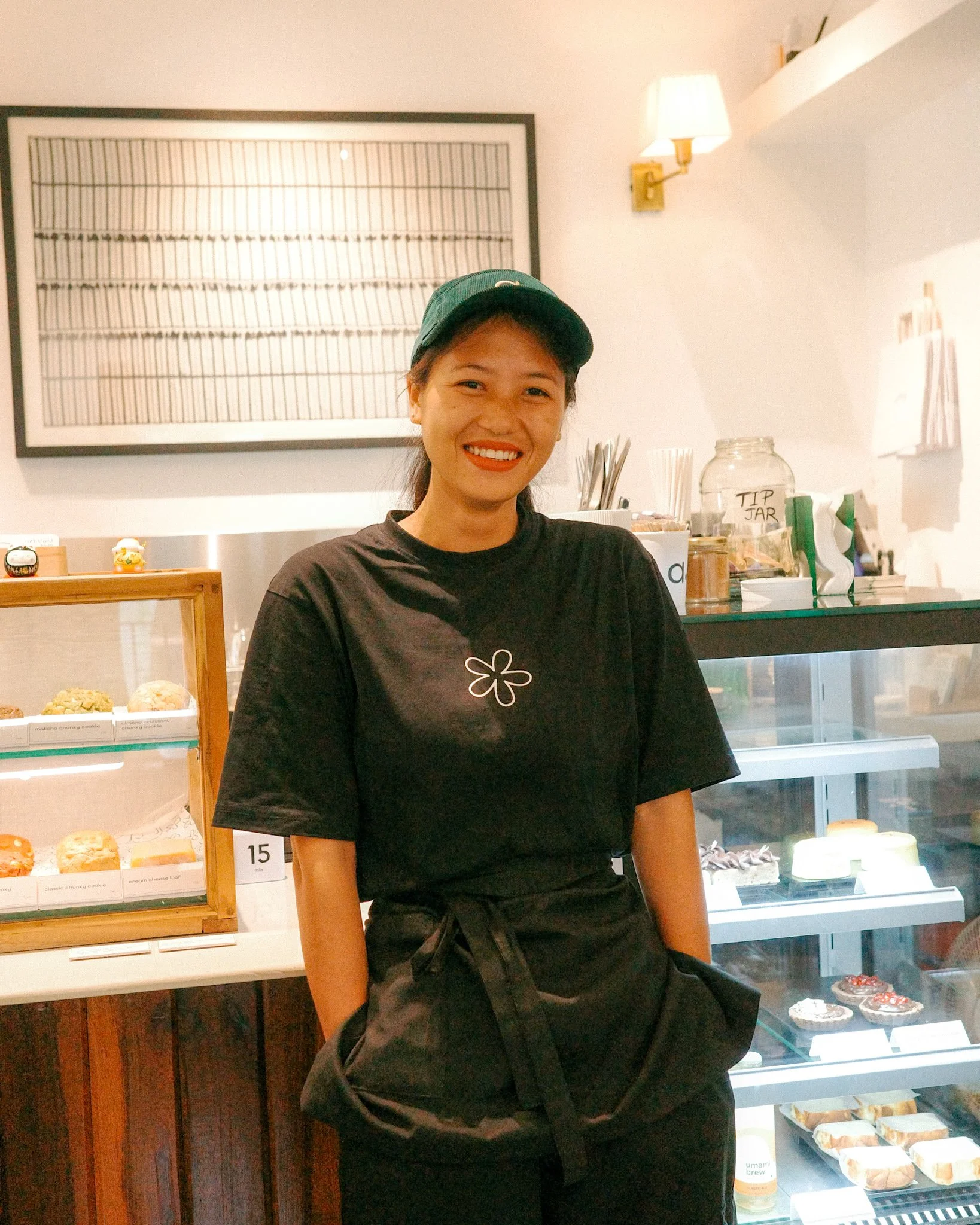 Smiling woman in black uniform and green cap standing behind bakery counter with pastries and desserts displayed.