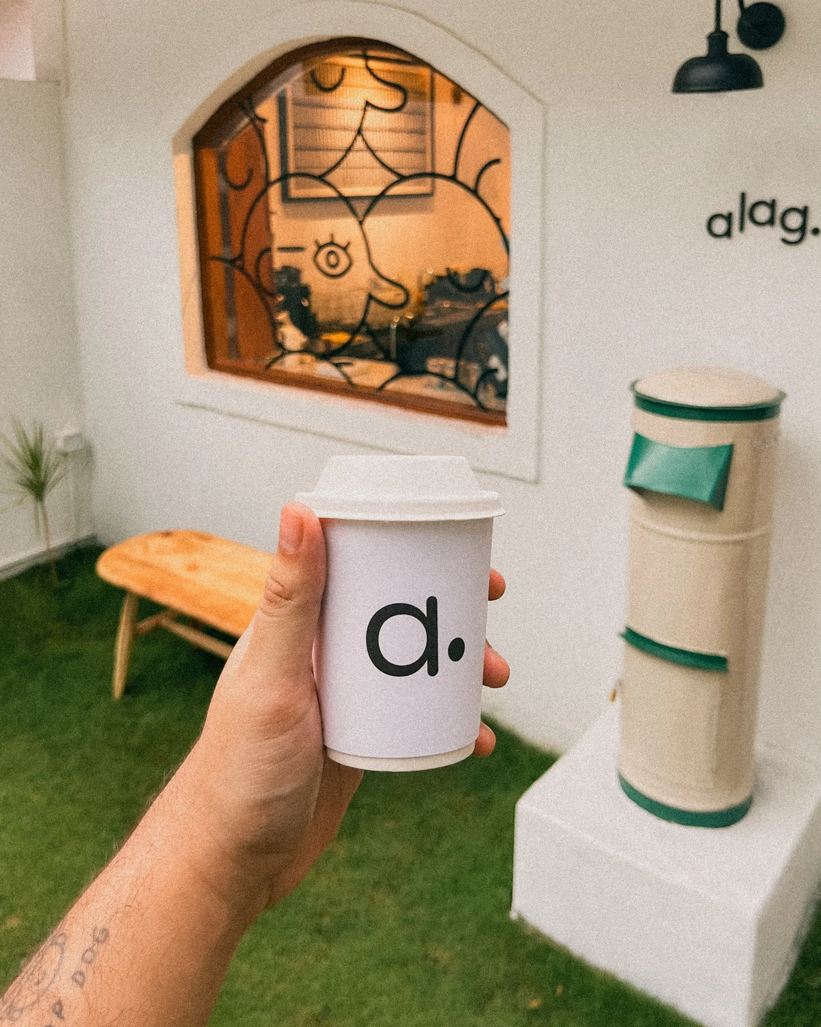 A person holds a white paper cup with the letter 'd.' printed on it. In the background, there is a green artificial grass floor, a small wooden bench, a white wall with a decorative window featuring black line art of a face, a small plant, and a beige and green mailbox.