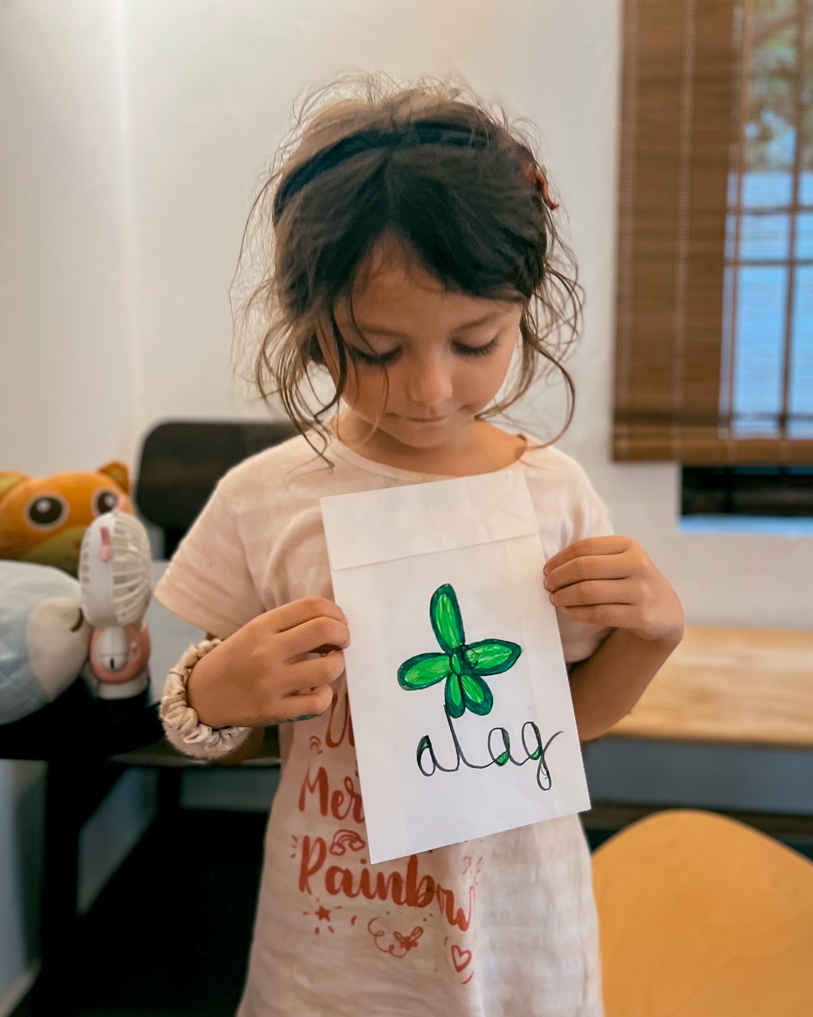 A young girl holding a drawing of a green four-leaf clover with the word 'aleg' written underneath. She has dark curly hair and is wearing a pale pink T-shirt.
