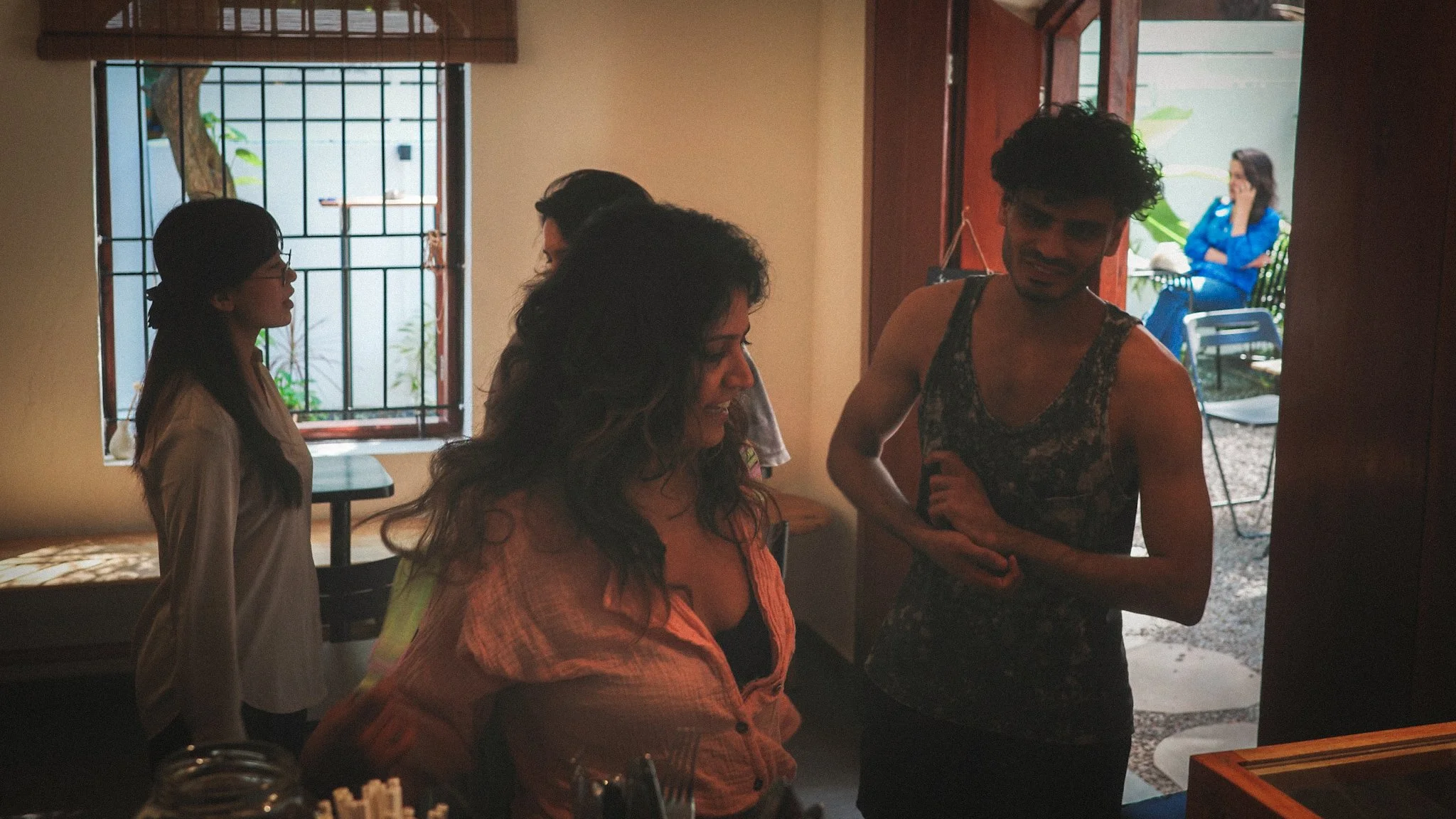 A group of four people inside a room with an open door showing a woman sitting outside. Two women and one man are standing and talking inside, while the woman outside is seated on a chair, on the phone.