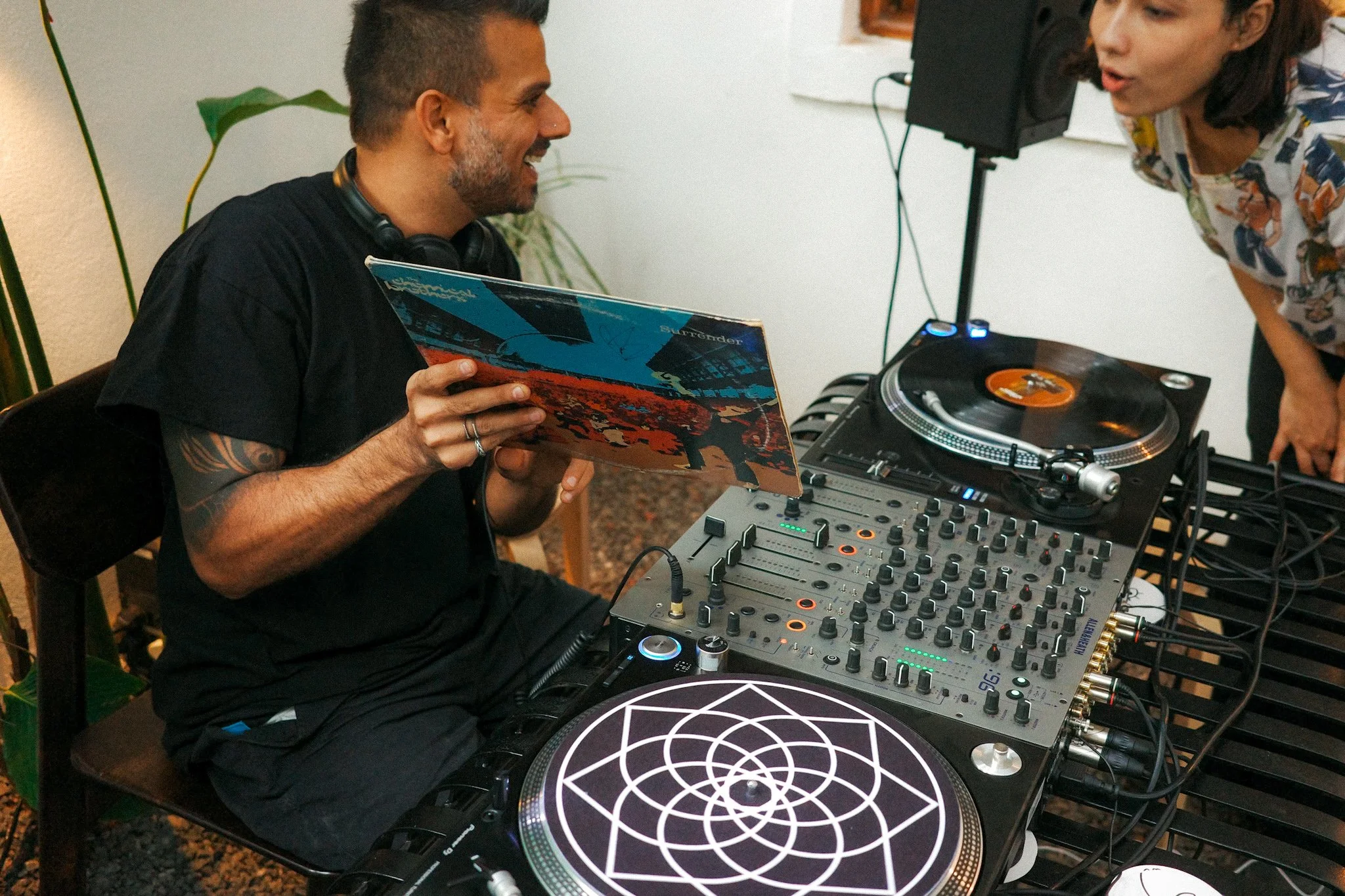 A man sitting with headphones around his neck, holding a vinyl record, smiling at a woman nearby, surrounded by DJ equipment, including turntables and a mixer, in an indoor setting.