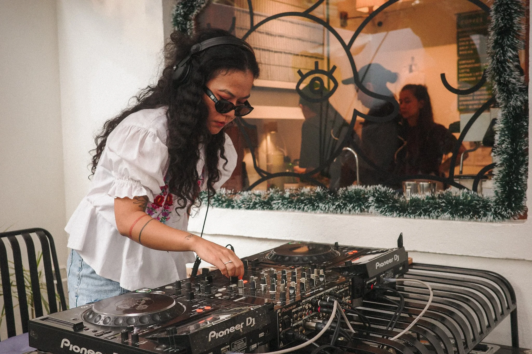 A woman with long curly hair, wearing sunglasses, white blouse, and light blue jeans, DJing with Pioneer DJ equipment in a decorated indoor setting, with Christmas wreaths and garlands, and people in the background through a window.