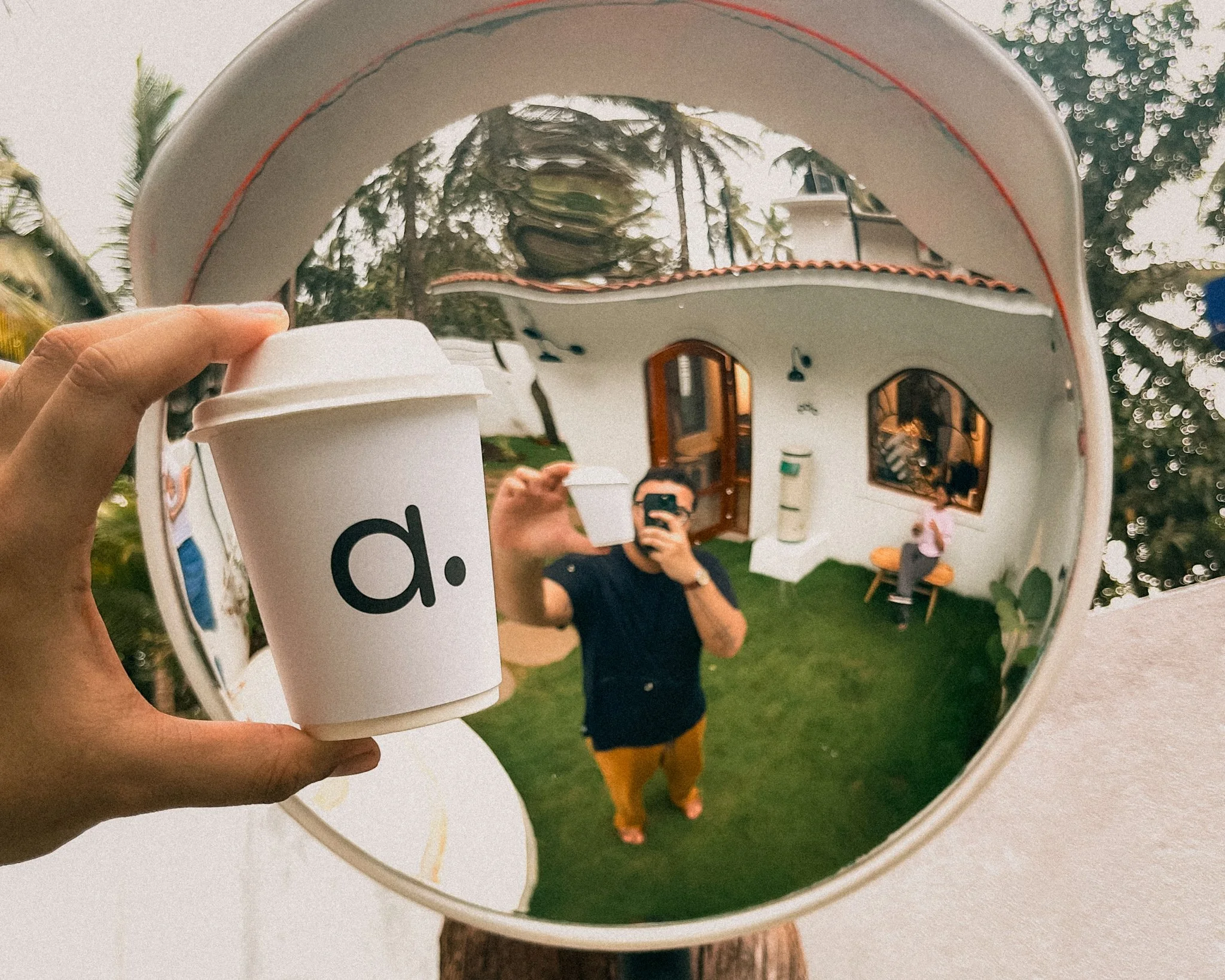 A person holding a white to-go coffee cup in front of a reflective, spherical surface, capturing a reflection of the person taking a photo with a smartphone, standing on grass outside a house with white walls, arched windows, and a wooden door, surro