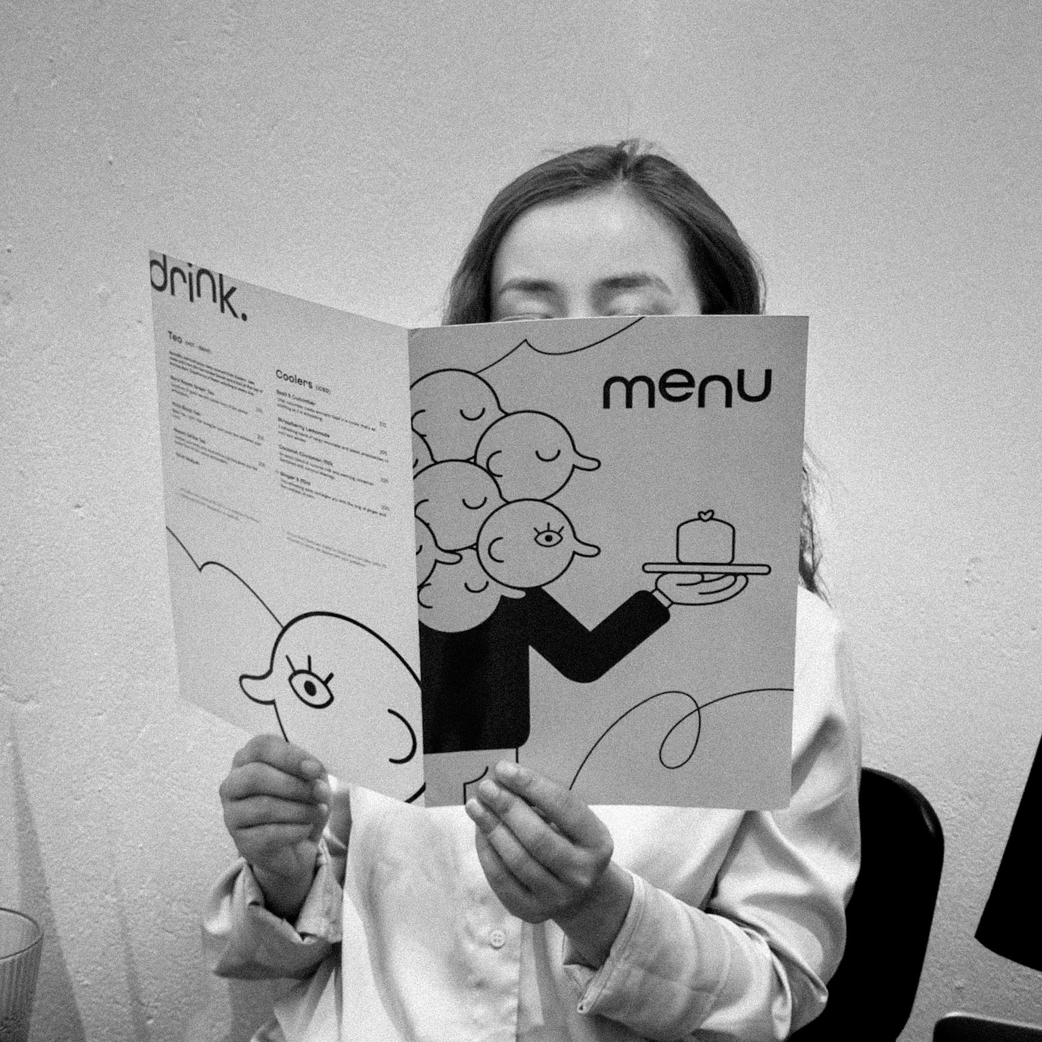 A person with long hair holding a colorful menu with cartoon illustrations of birds and a hand with a dessert, while sitting against a plain wall.