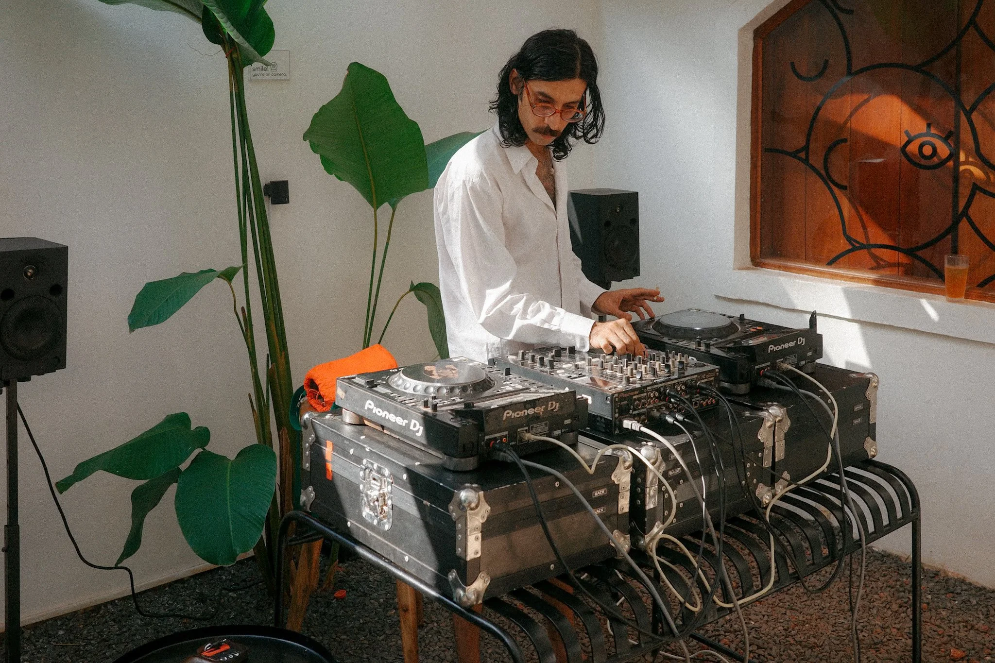 A man with dark wavy hair, glasses, and a mustache, wearing a white shirt, DJing with Pioneer equipment at an indoor venue. There are large green plants, speakers, and a decorative window in the background.