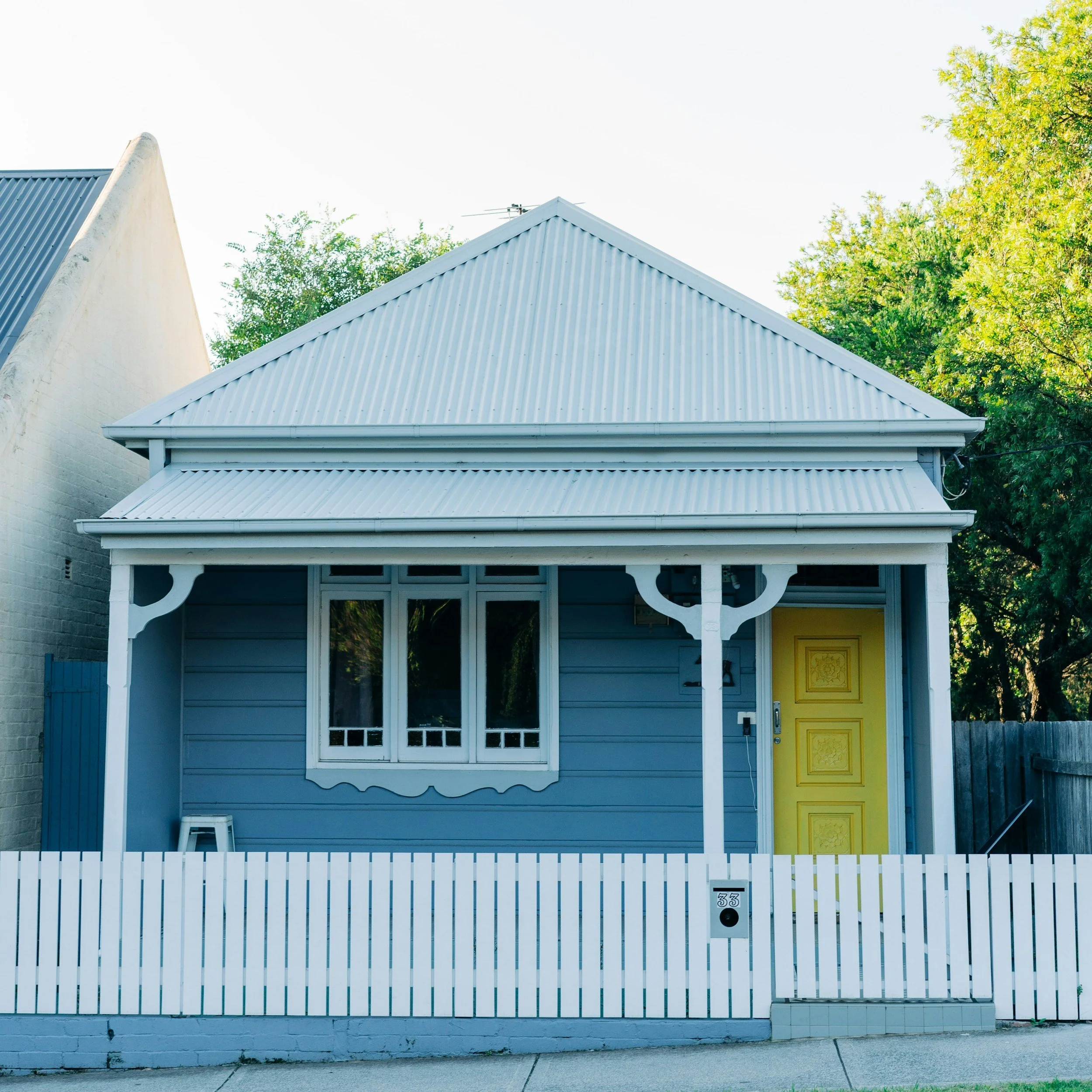 Front view of a light blue house with a corrugated metal roof, white trim, and a white picket fence.