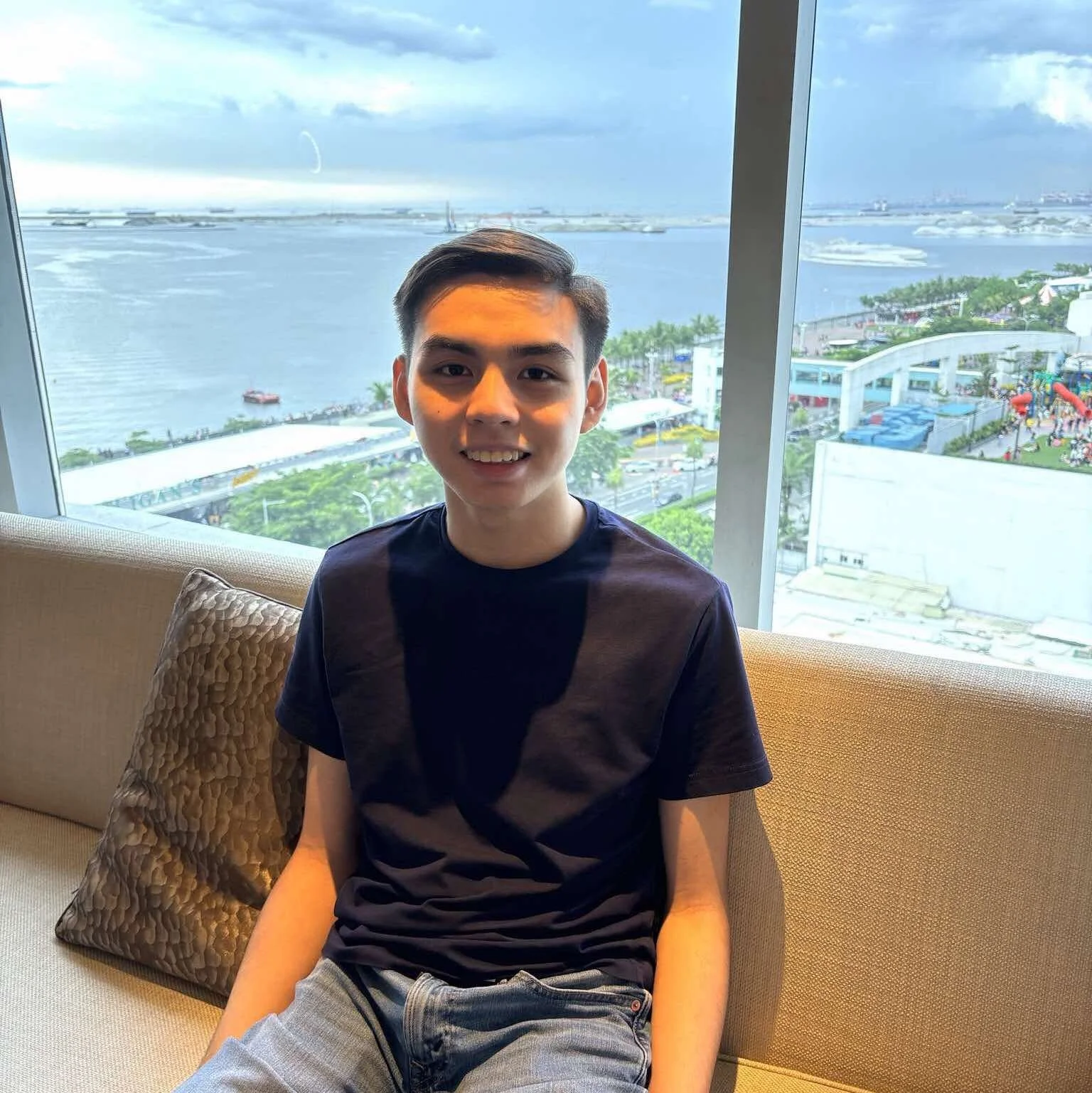 A young man sitting on a beige couch with a patterned pillow, smiling, with large windows overlooking a modern waterfront cityscape with water, trees, and buildings in the background.