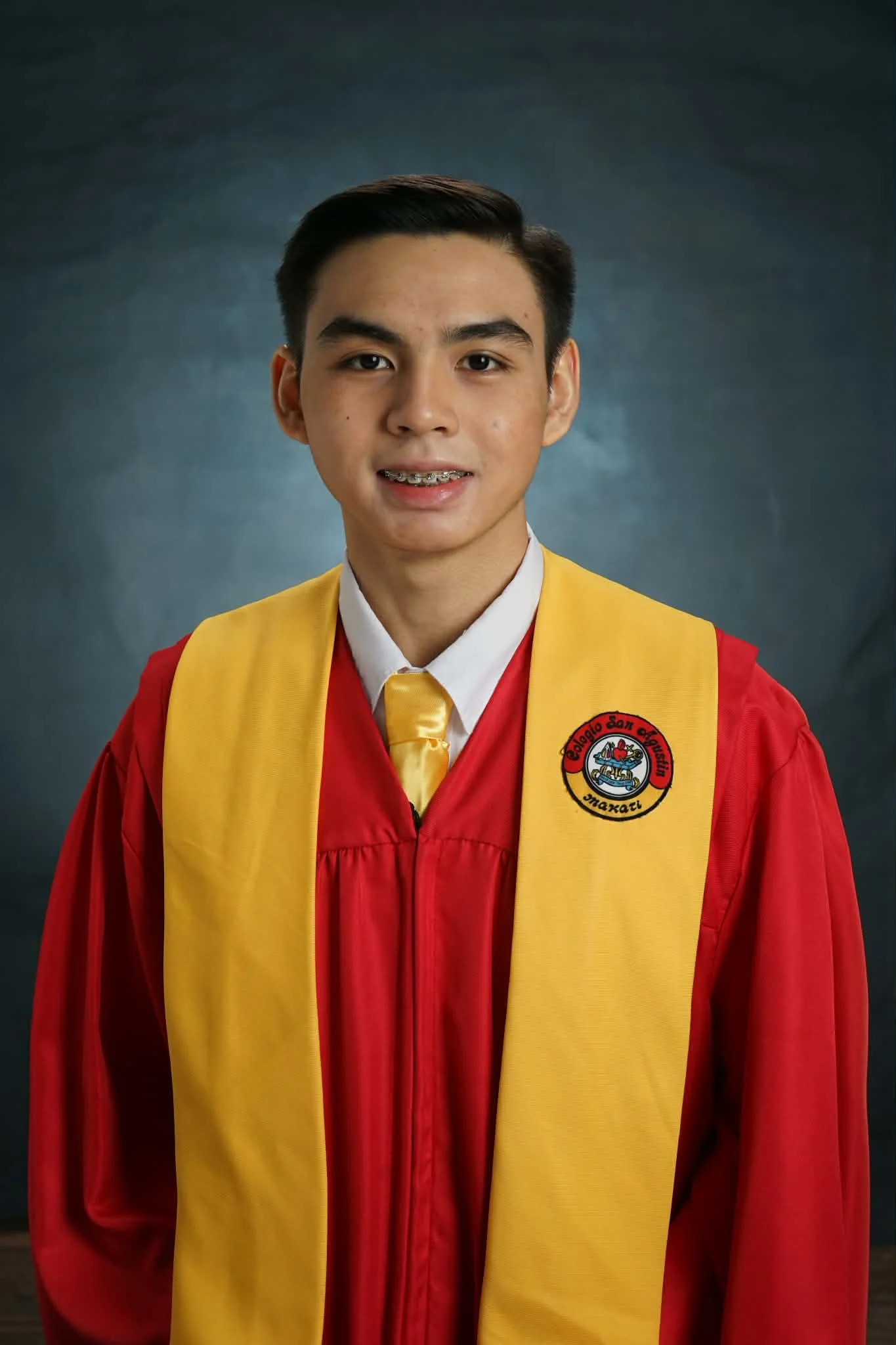 A young man in a red graduation gown with a yellow stole and a yellow tie, smiling for a formal portrait against a dark gray background.
