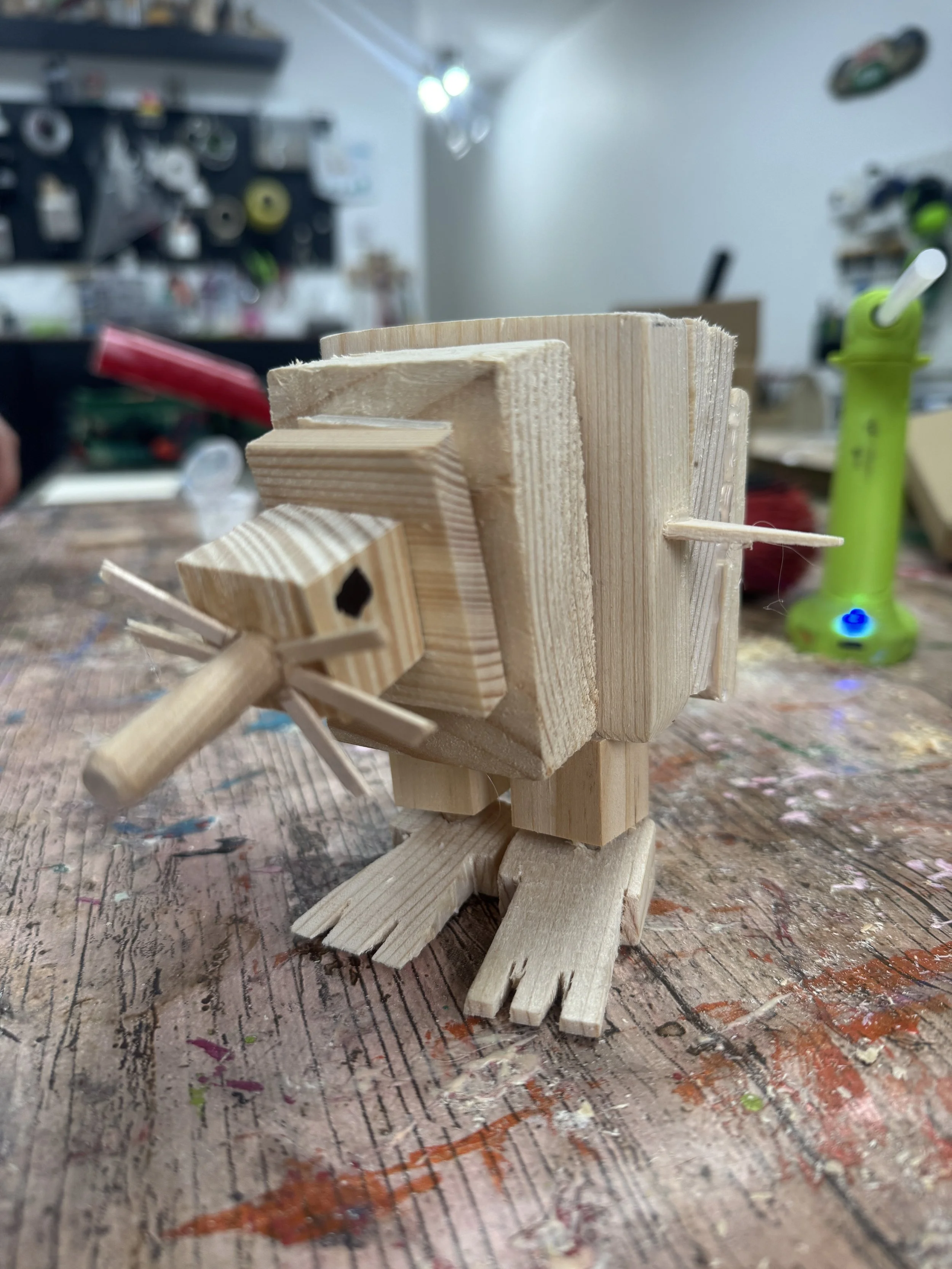 A small wooden sculpture resembling a bird or animal, constructed from various cut wood pieces, is displayed on a workbench in a workshop.