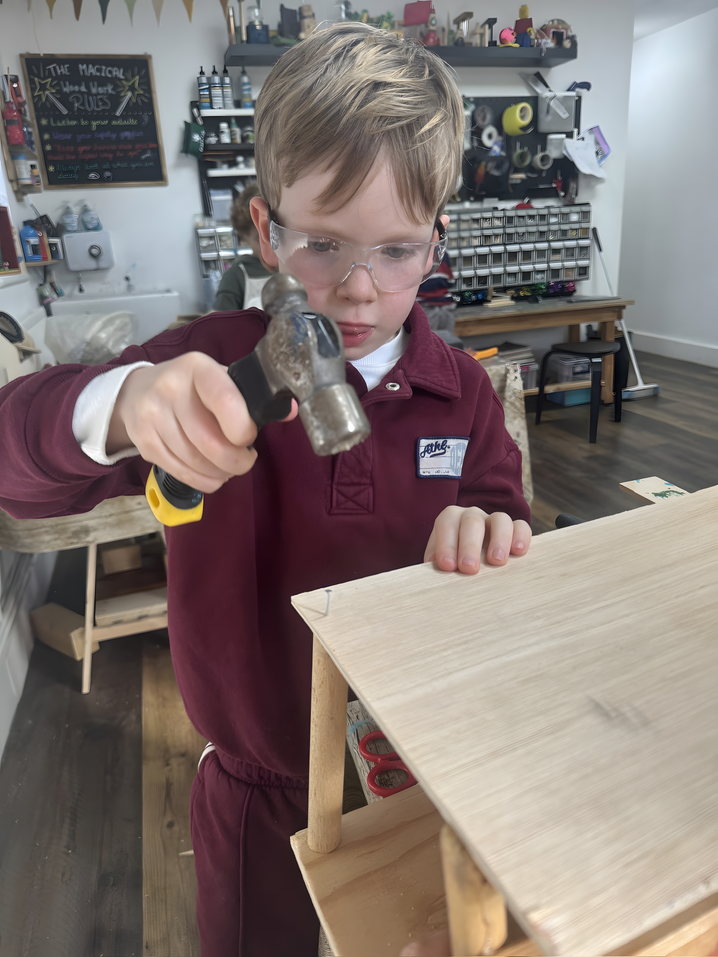 A young boy wearing safety glasses and a burgundy uniform hammering a nail into a wooden project in a workshop or craft room.