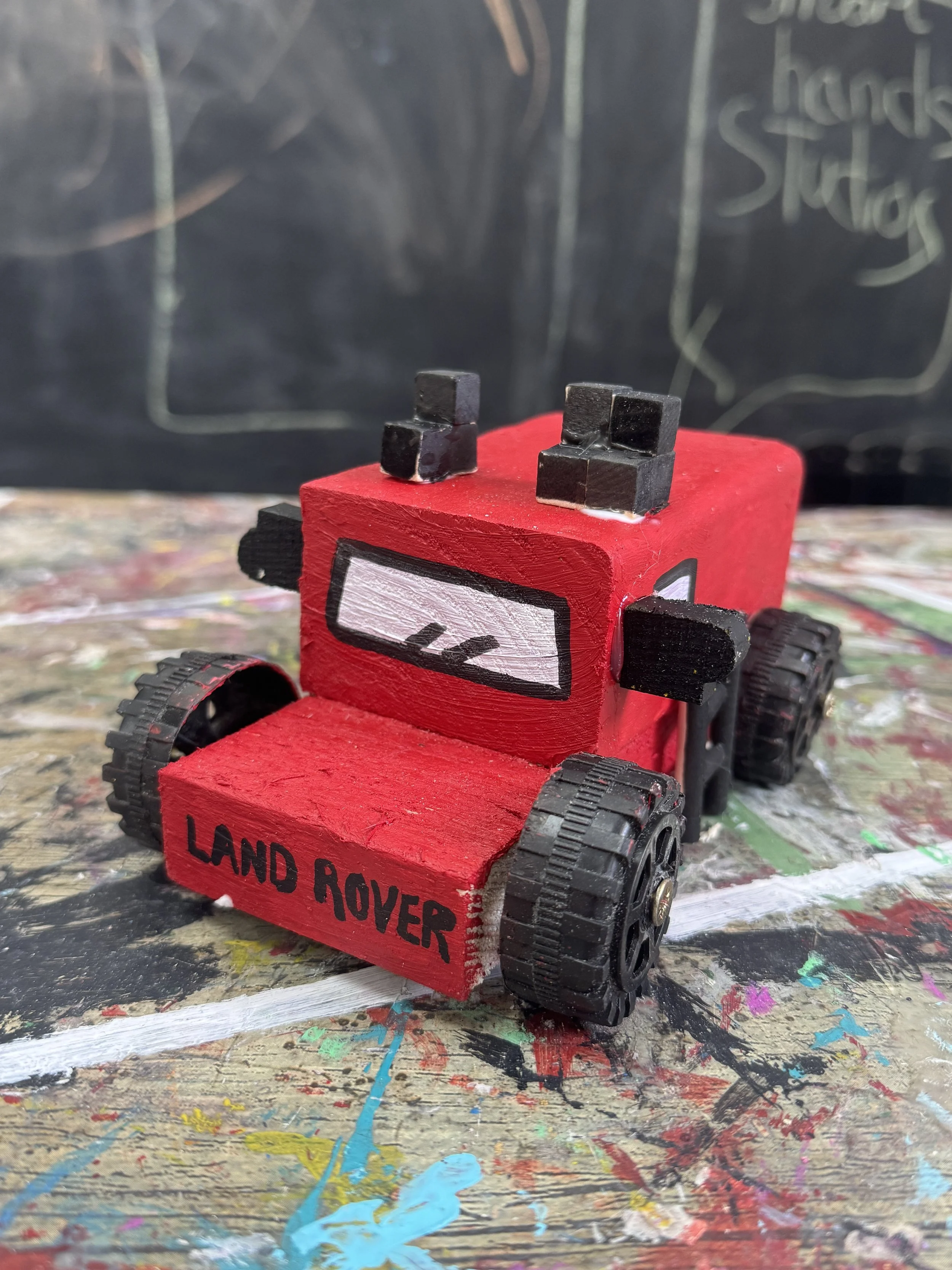A small red wooden toy vehicle resembling a Land Rover, with painted black wheels, black side mirrors, and a cartoonish face on the front. The words 'LAND ROVER' are written on the front bumper. The toy is set on a painted, colorful surface with a bl