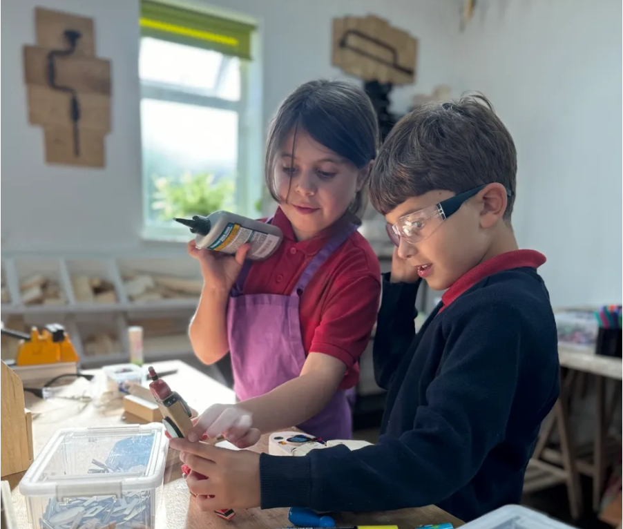 Two children working together on a woodworking project in a workshop, using tools and safety glasses.
