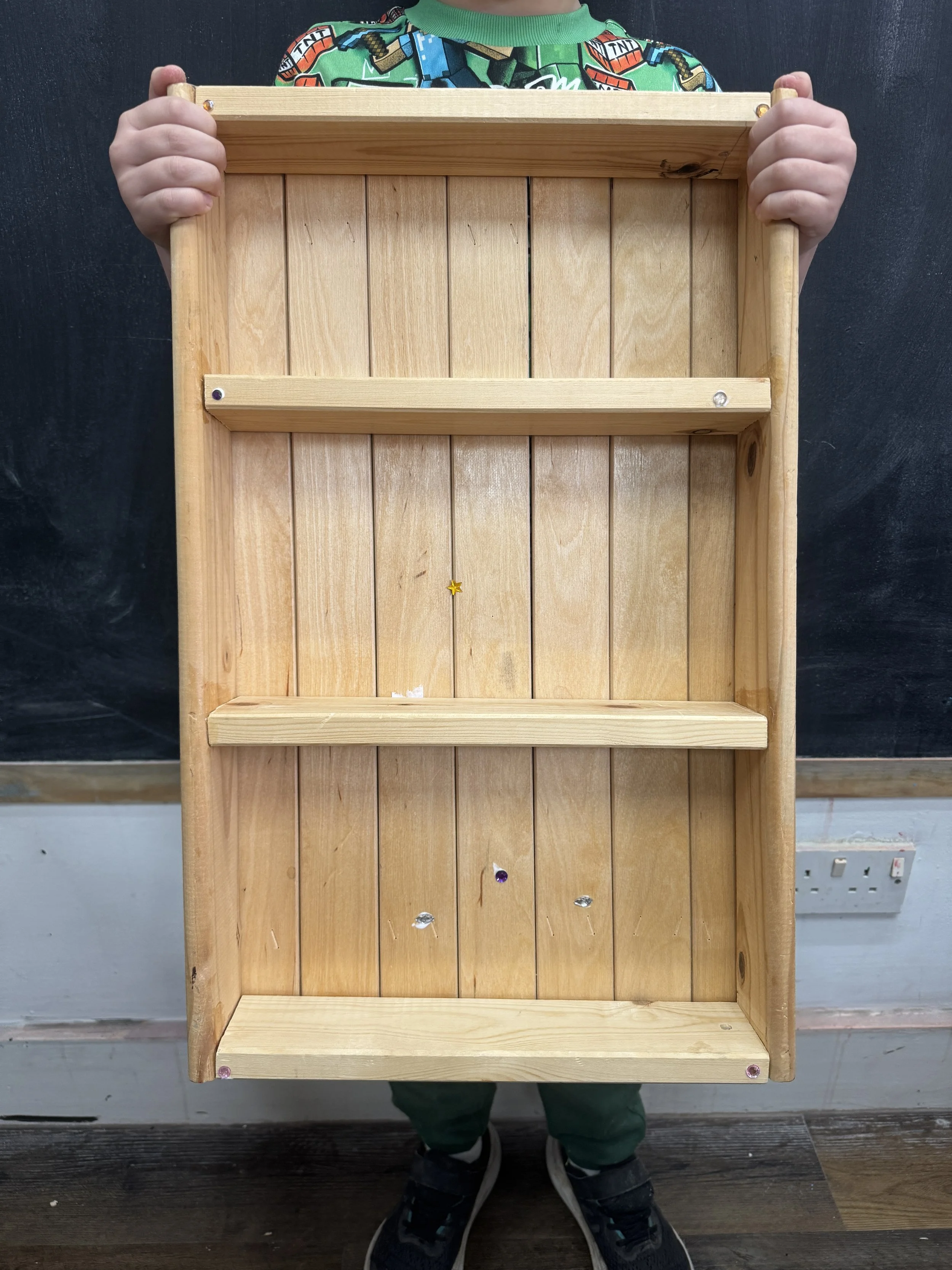 Child holding a wooden shelf with three levels, no items on the shelf, in front of a blackboard and electrical outlets on the wall.