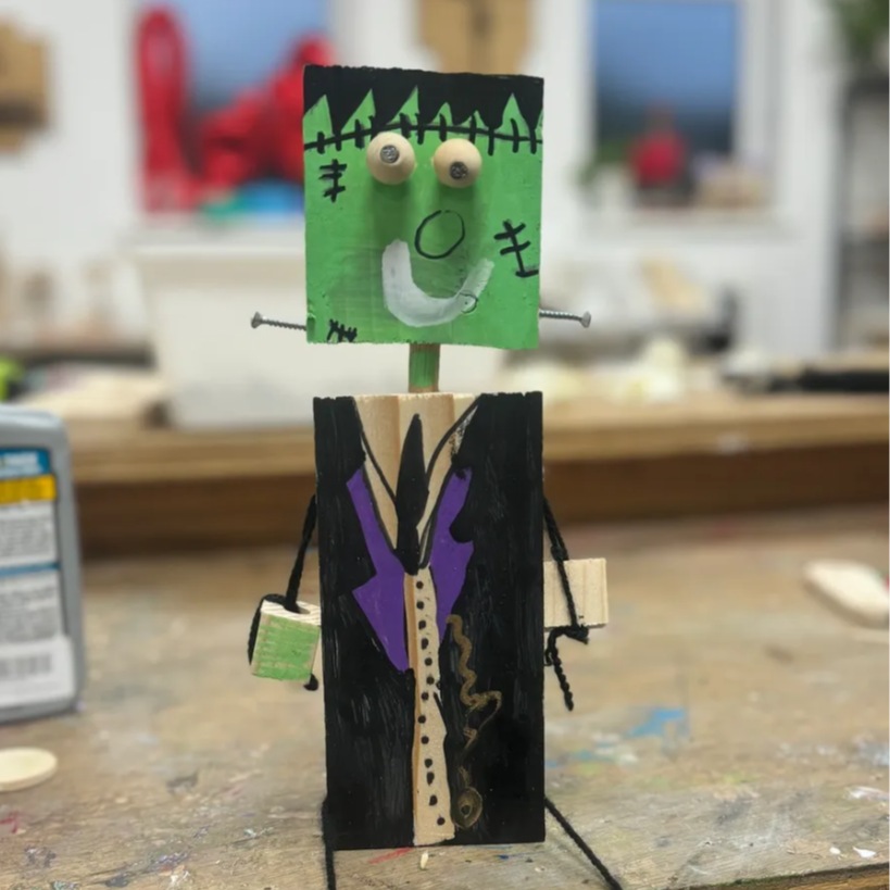 A homemade robot sculpture with a green Frankenstein face made of painted wood, with googly eyes, a drew smile, and stitches, mounted on a wooden stick. The body is painted black with a purple vest and a beige shirt, with wire arms and small wooden b
