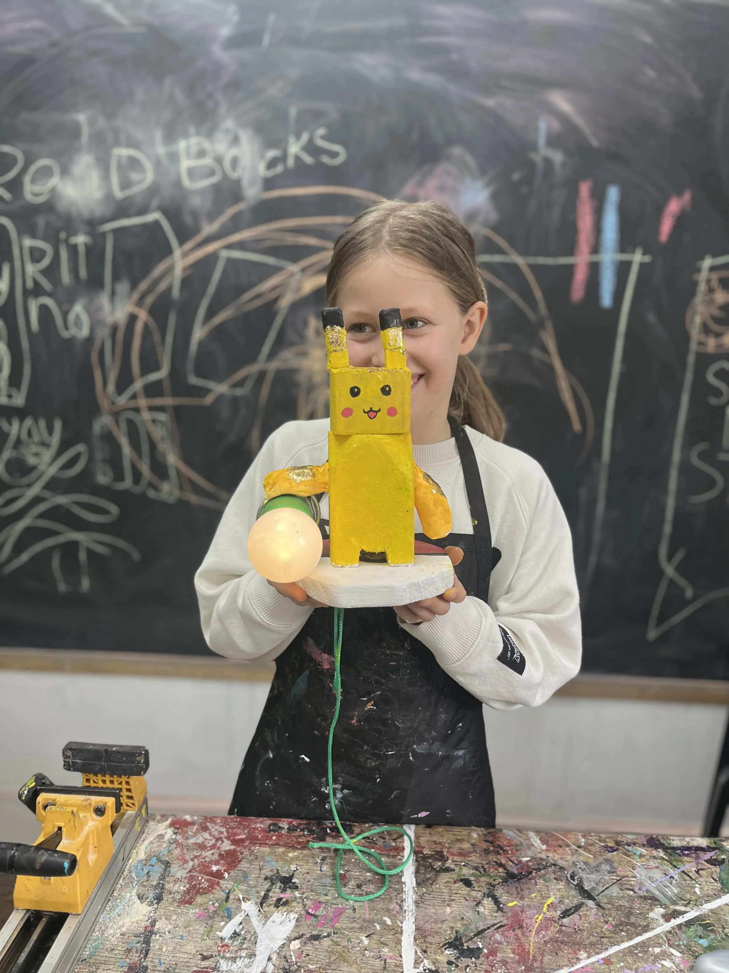 A young girl holding a handmade Pikachu craft with a glowing orb, standing in front of a chalkboard with wall scribbles. She is smiling and wearing an apron.