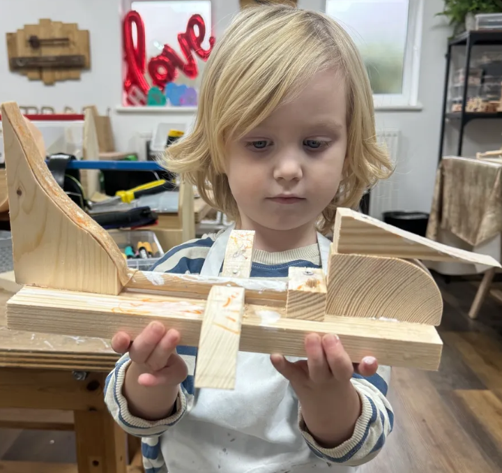 A young boy with blond hair examines a wooden winged craft project in a workshop, with various tools and shelves in the background.