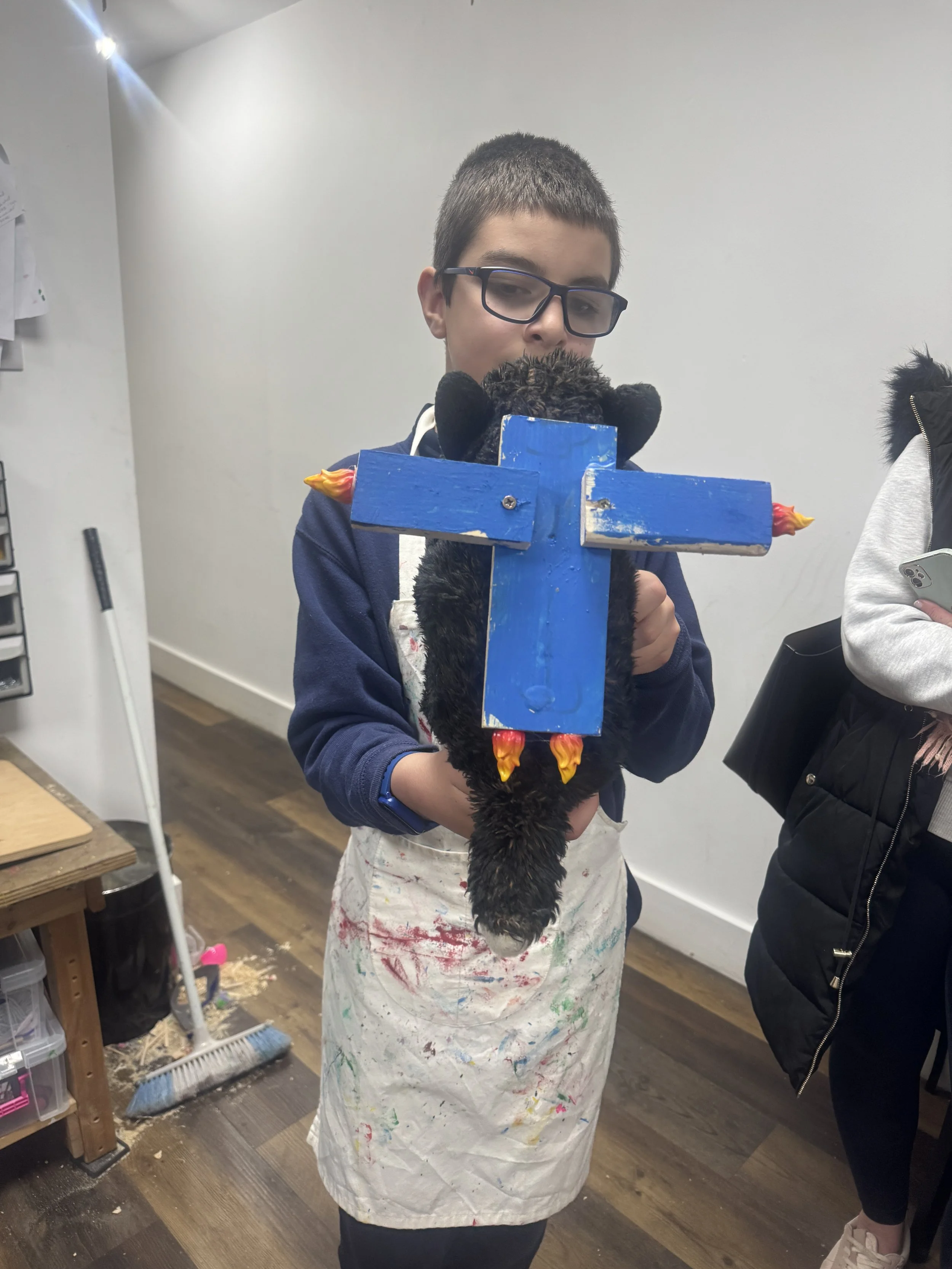 A boy with glasses wearing an apron holding a black plush pig toy with a blue wooden cross mounted on its back, with small flame-shaped details at the ends.