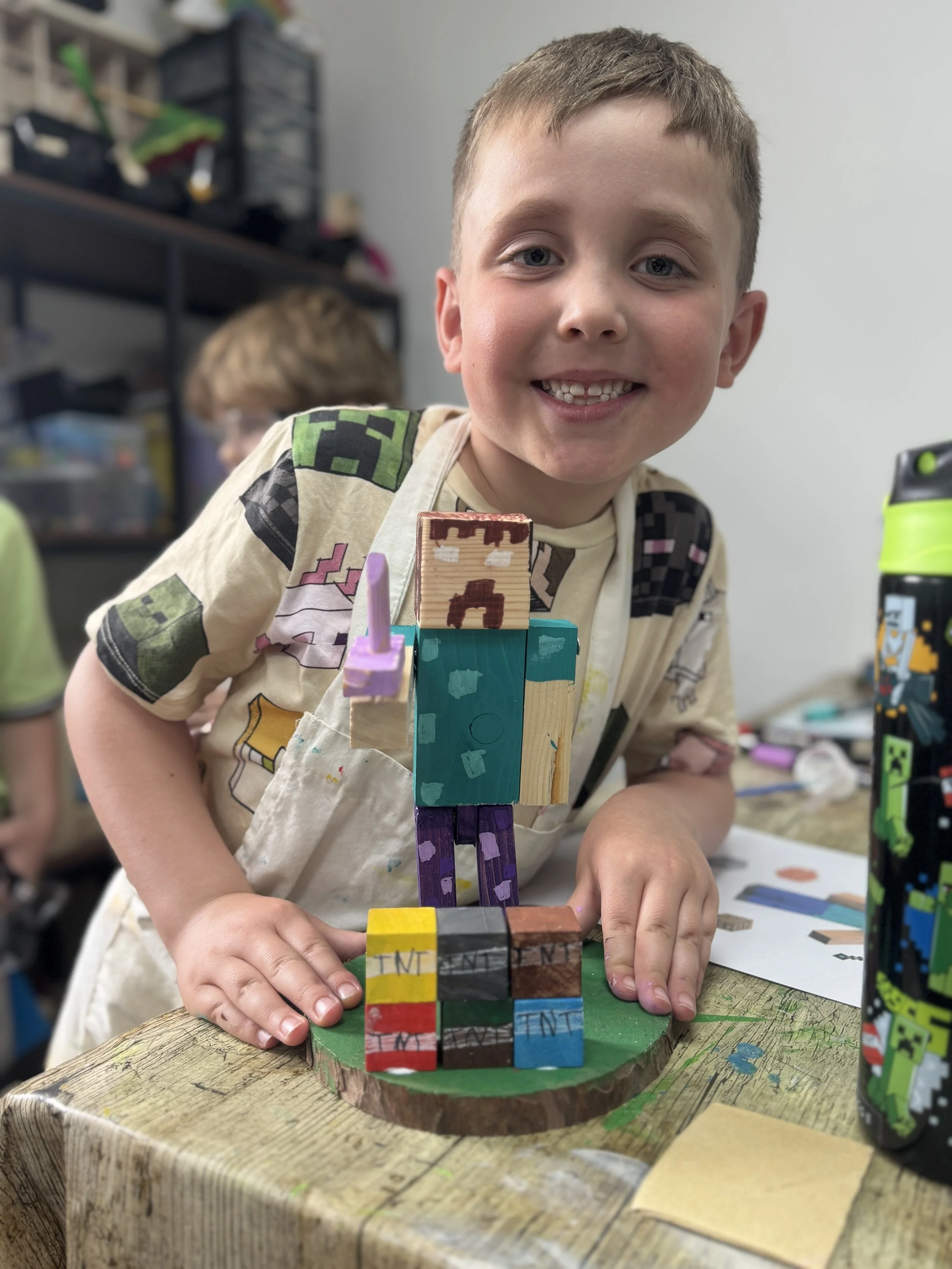 A young boy smiling and showing a Minecraft-themed craft project made from decorated wooden blocks on a circular wooden base, with a building block figure on top, in a classroom setting.