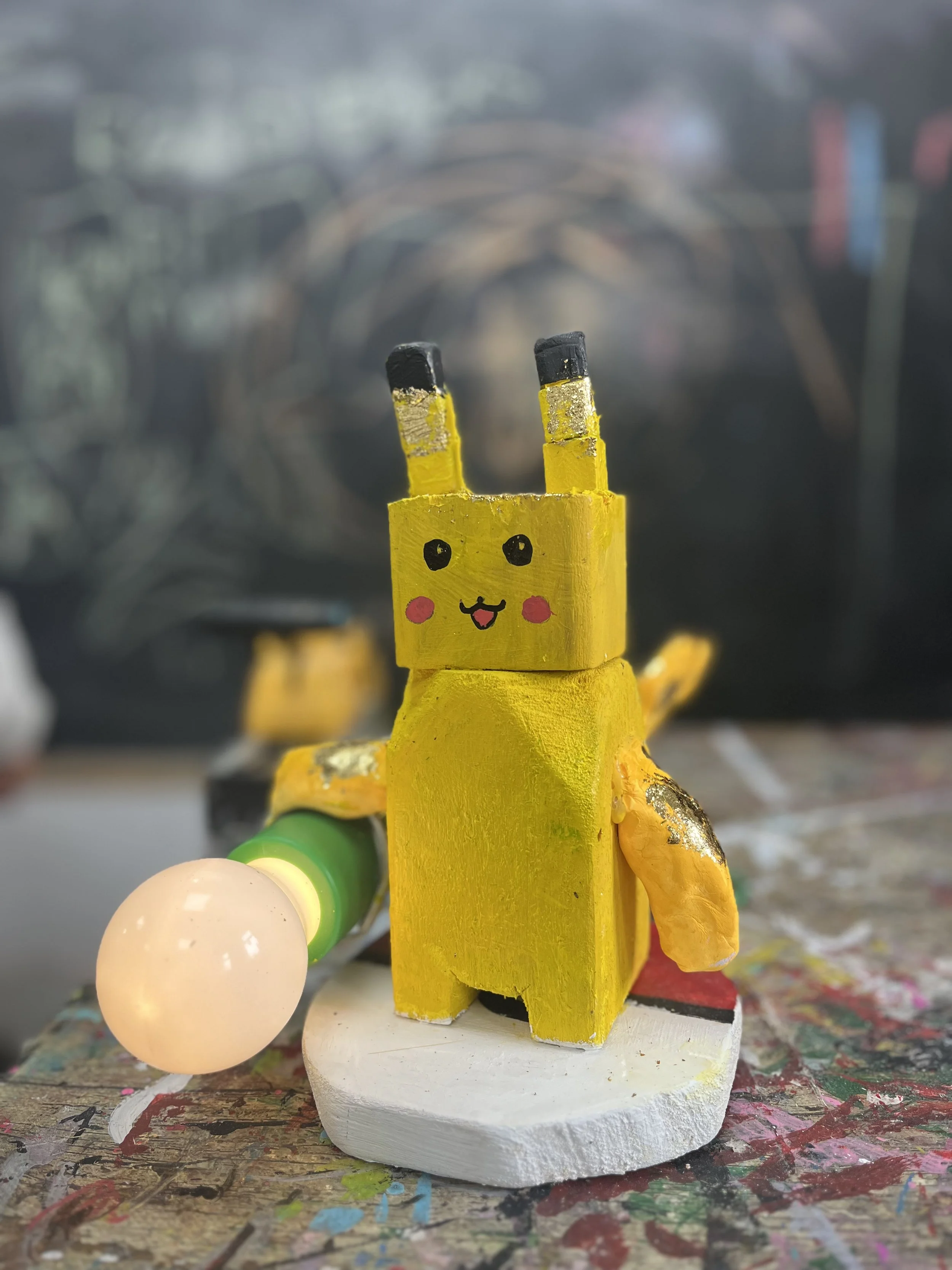 Handmade wooden robot figure painted yellow with black-tipped antennae, pink cheeks, and a smiling face, holding a glowing orb in a workshop setting.
