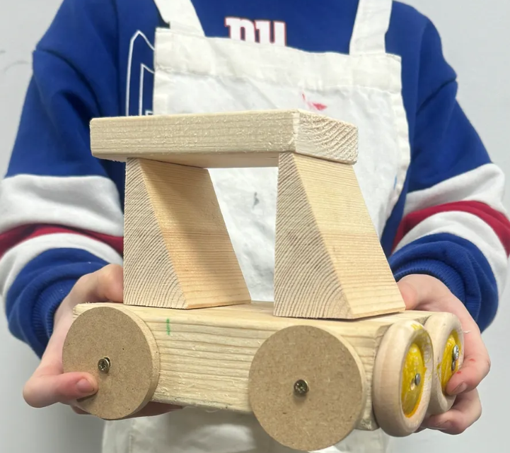 Child holding a wooden toy car with circular wheels and a cabin top, wearing a blue and white sports jersey.