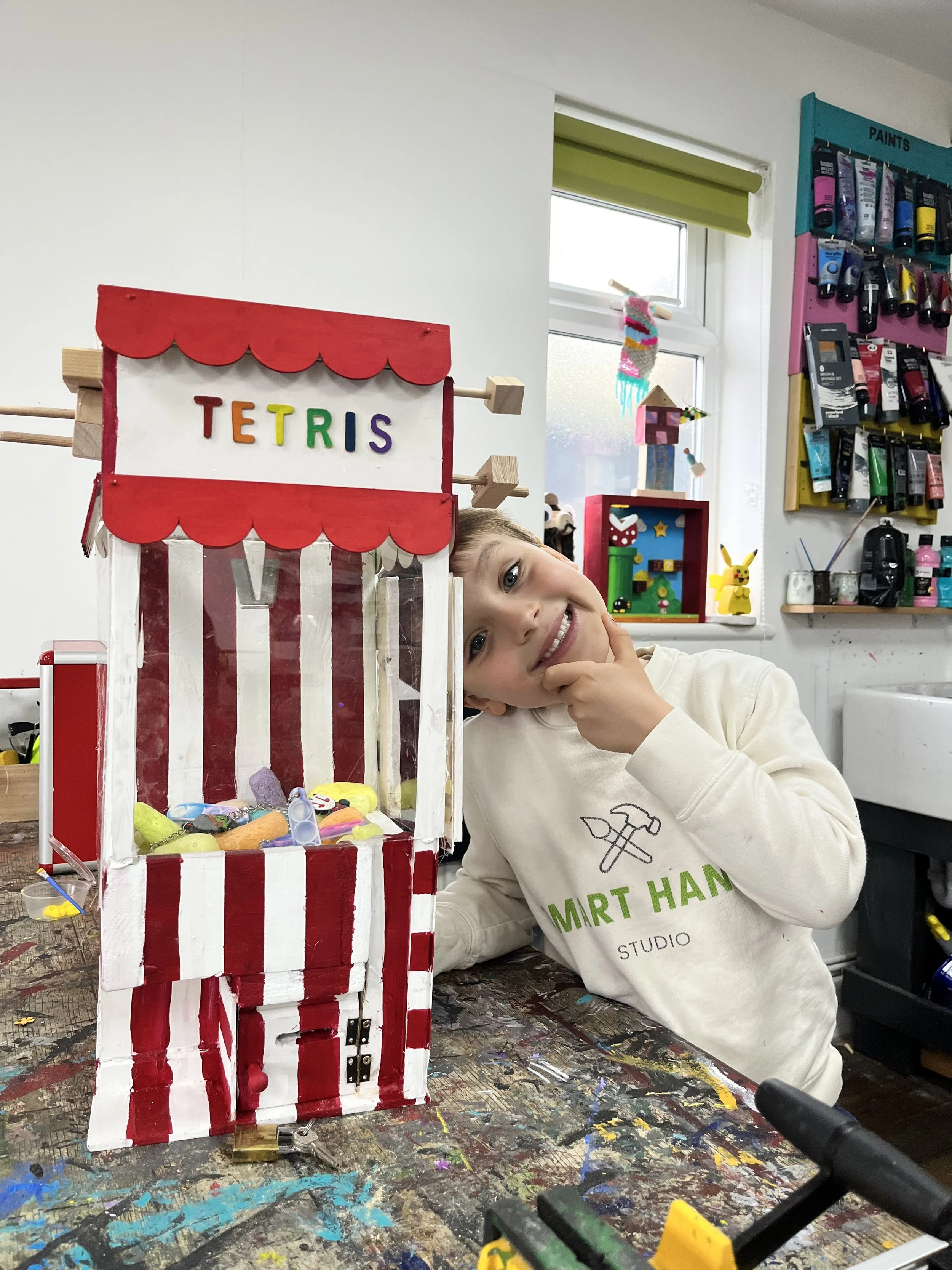 A young boy smiles and leans his head against a colorful homemade Tetris arcade game display, with a workshop or art room background.