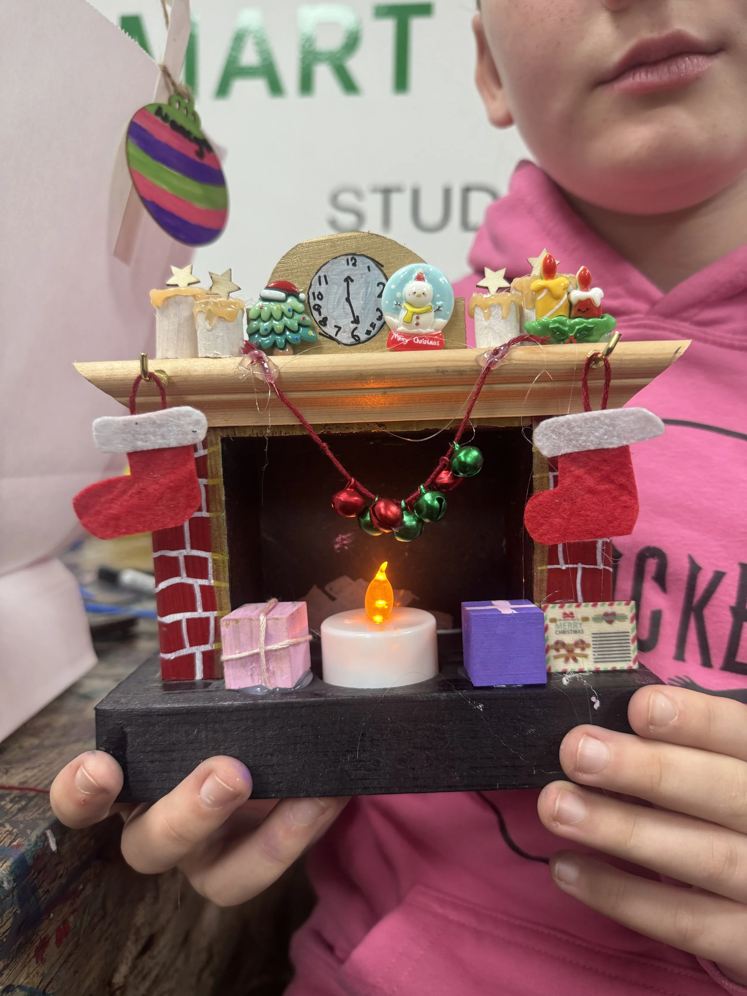 Child holding a Christmas-themed miniature house decoration with candles, gifts, and festive ornaments.
