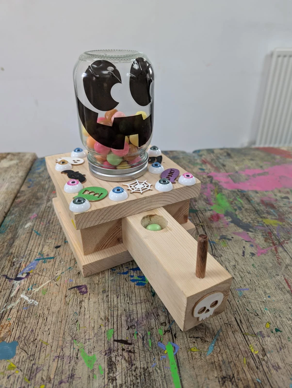 A Halloween-themed craft project with a wooden base, decorated with googly eyes, small skull and spider accessories, and a glass jar painted with a face, filled with colorful candy and small toys, set on a wooden table with paint splatters.