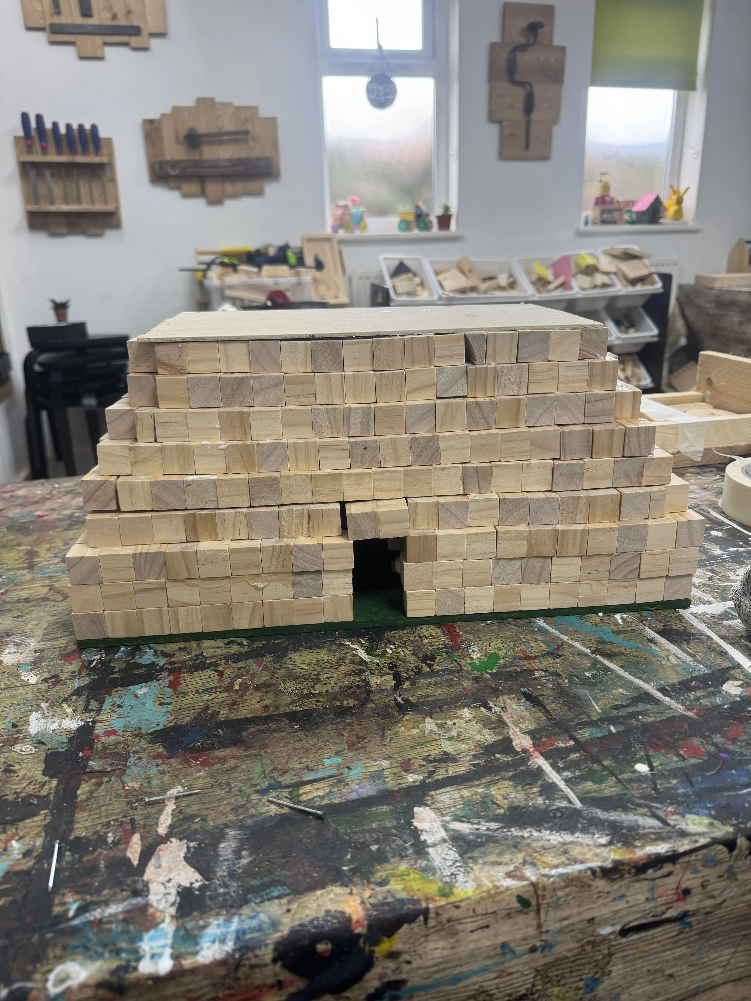 A miniature wooden house model under construction on a paint-splattered worktable inside a woodworking workshop.