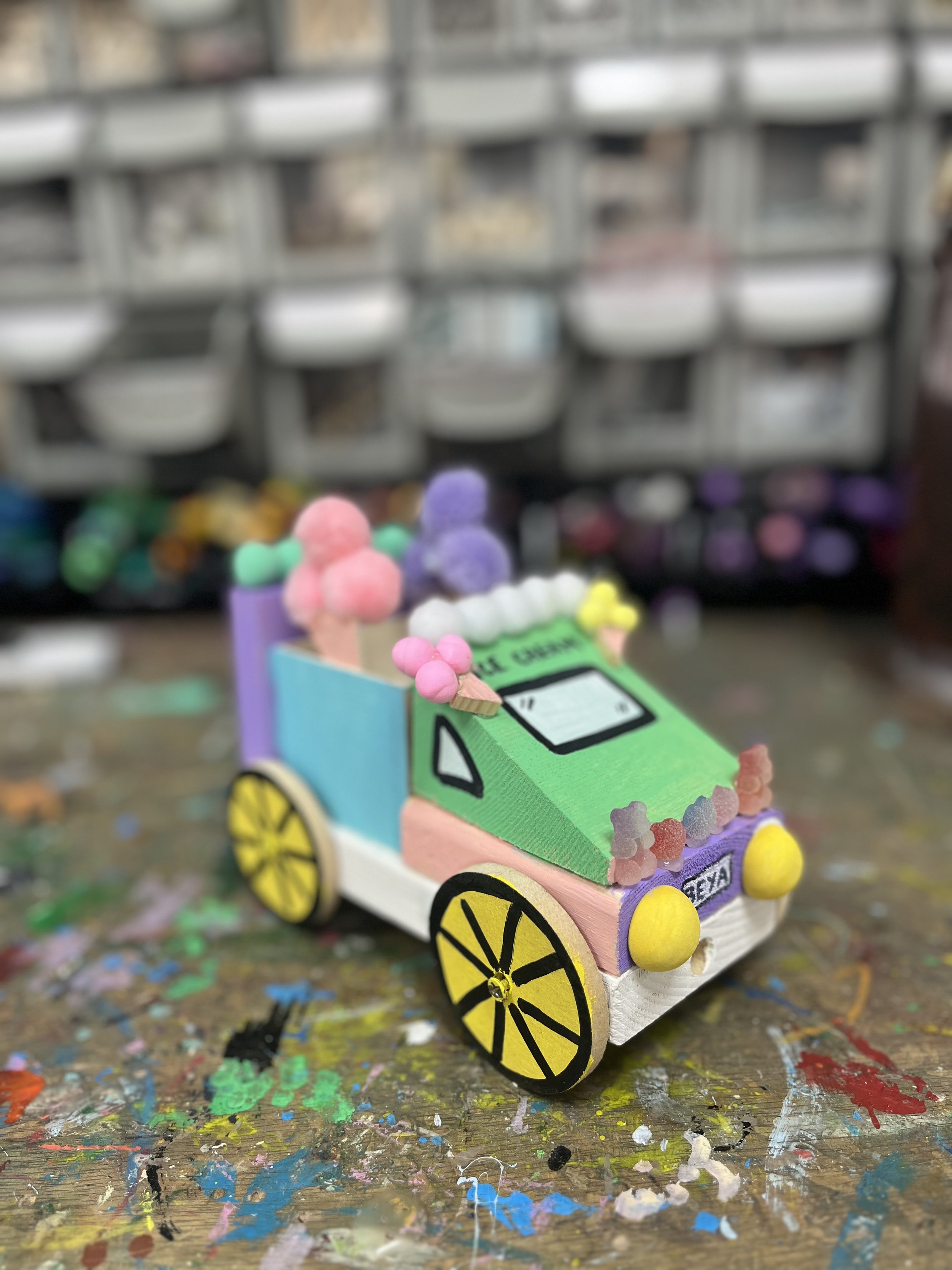 A colorful, handcrafted model of a cartoonish car with pastel colors and decorative elements, placed on a paint-splattered surface.