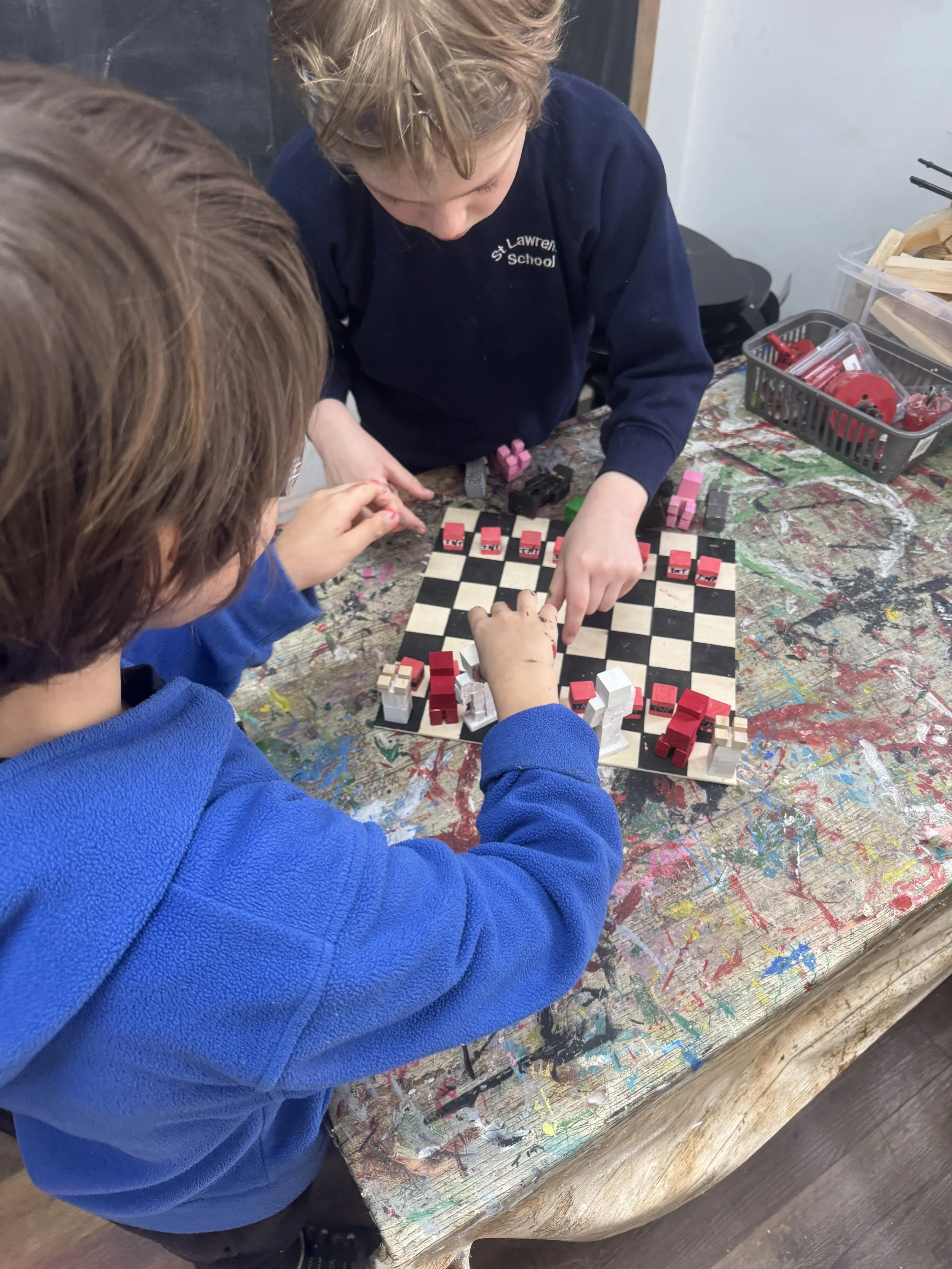 Two children playing chess with wooden pieces at a colorful, paint-splattered table.