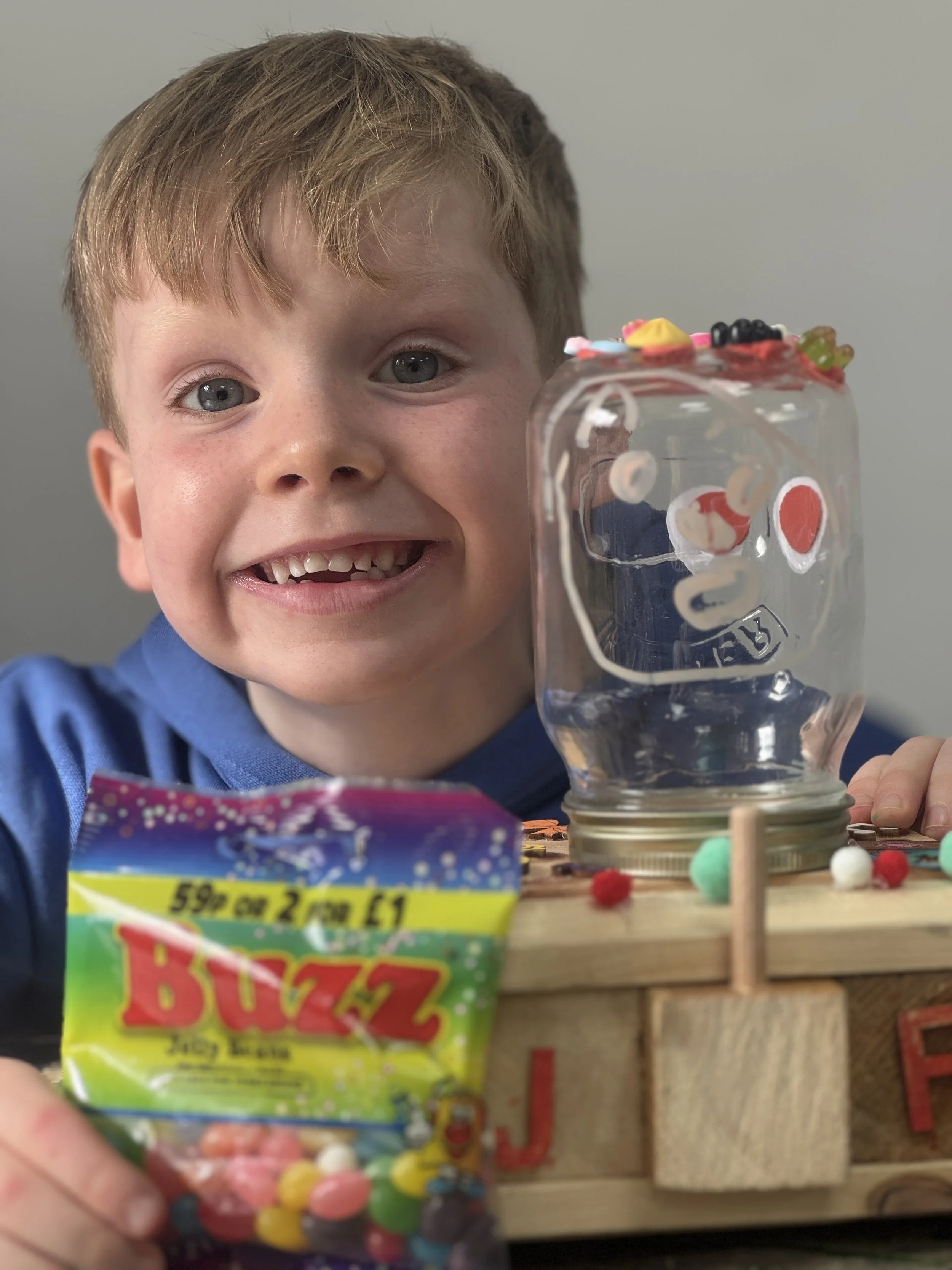 A young boy with light brown hair and blue eyes smiling at the camera, holding a small open bag of colorful candy in front of a makeshift craft display.