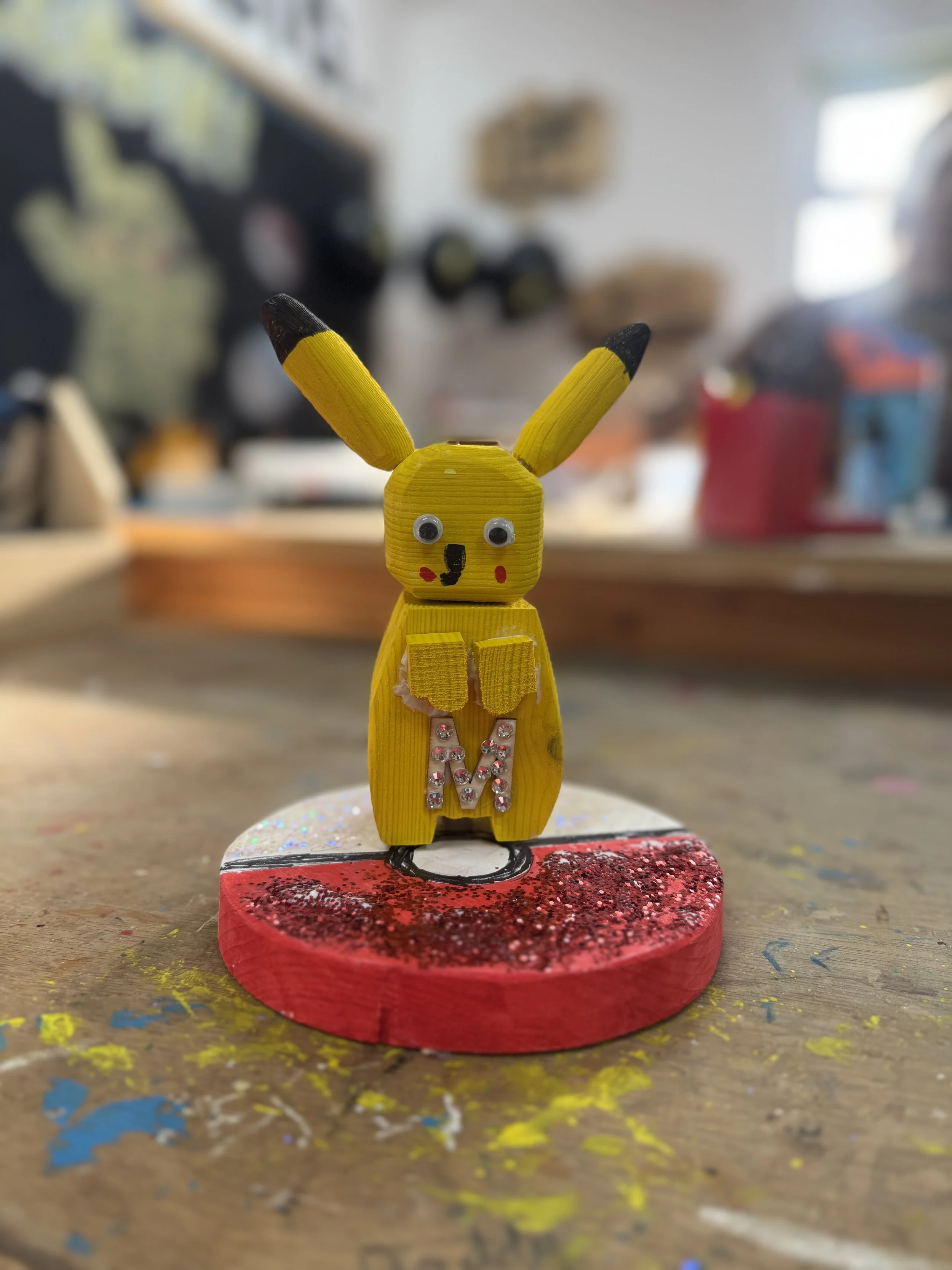A yellow Pikachu figurine with googly eyes and a painted face, holding a glittery pink letter M, stands on a red, glittery base on a wooden table with colorful paint splatters in the background.