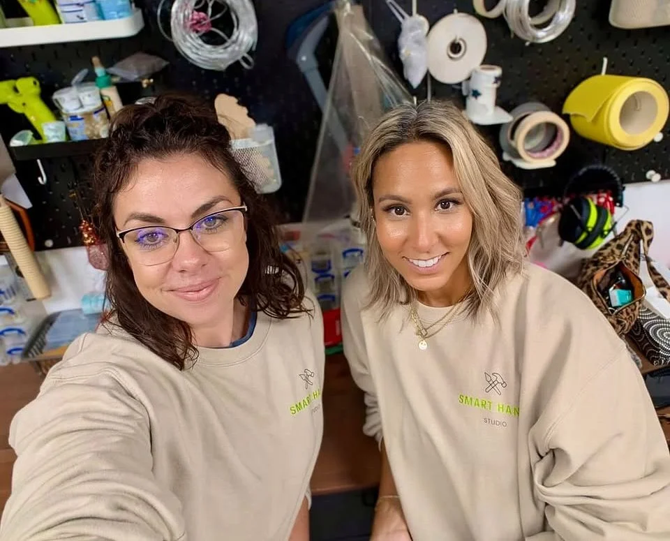 Two women in beige shirts with 'SMART HAND' on the front, smiling in a workshop with tools, tapes, and crafting supplies behind them.