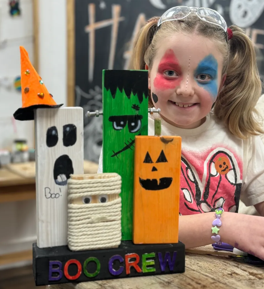 A smiling young girl with face paint resembling a clown, wearing safety glasses and a bunny fairy-tale themed shirt, is sitting at a wooden table with decorated Halloween-themed wooden blocks in front of her. The blocks are painted and decorated as a