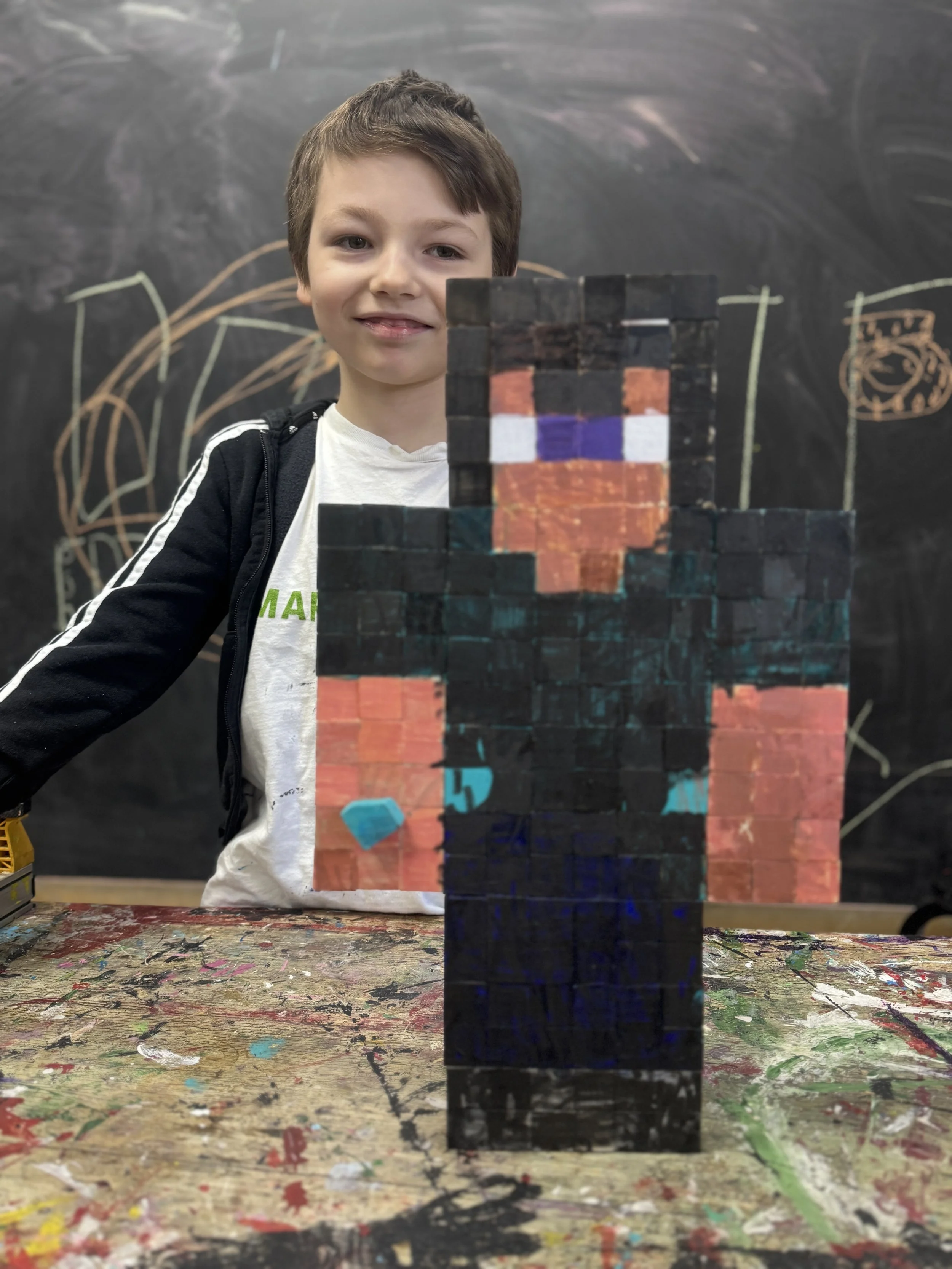 A young boy with brown hair smiling and standing behind a sculpture of a character from Minecraft, made of painted wooden blocks, in front of a black chalkboard with colorful chalk drawings.