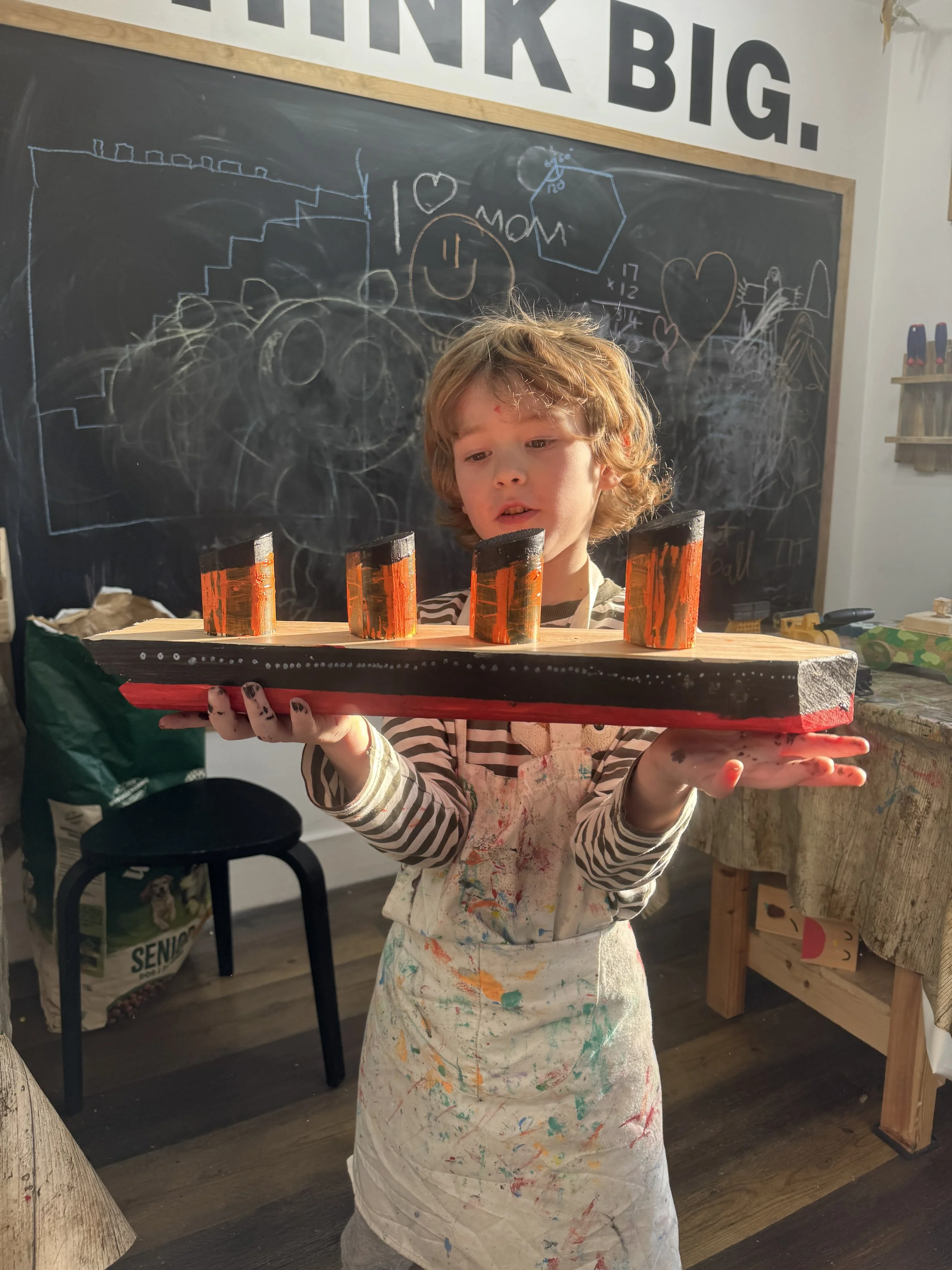 Child holding a handmade wooden boat model painted black and orange, covered in paint splatters, in a classroom with a blackboard and chalk drawings in the background.