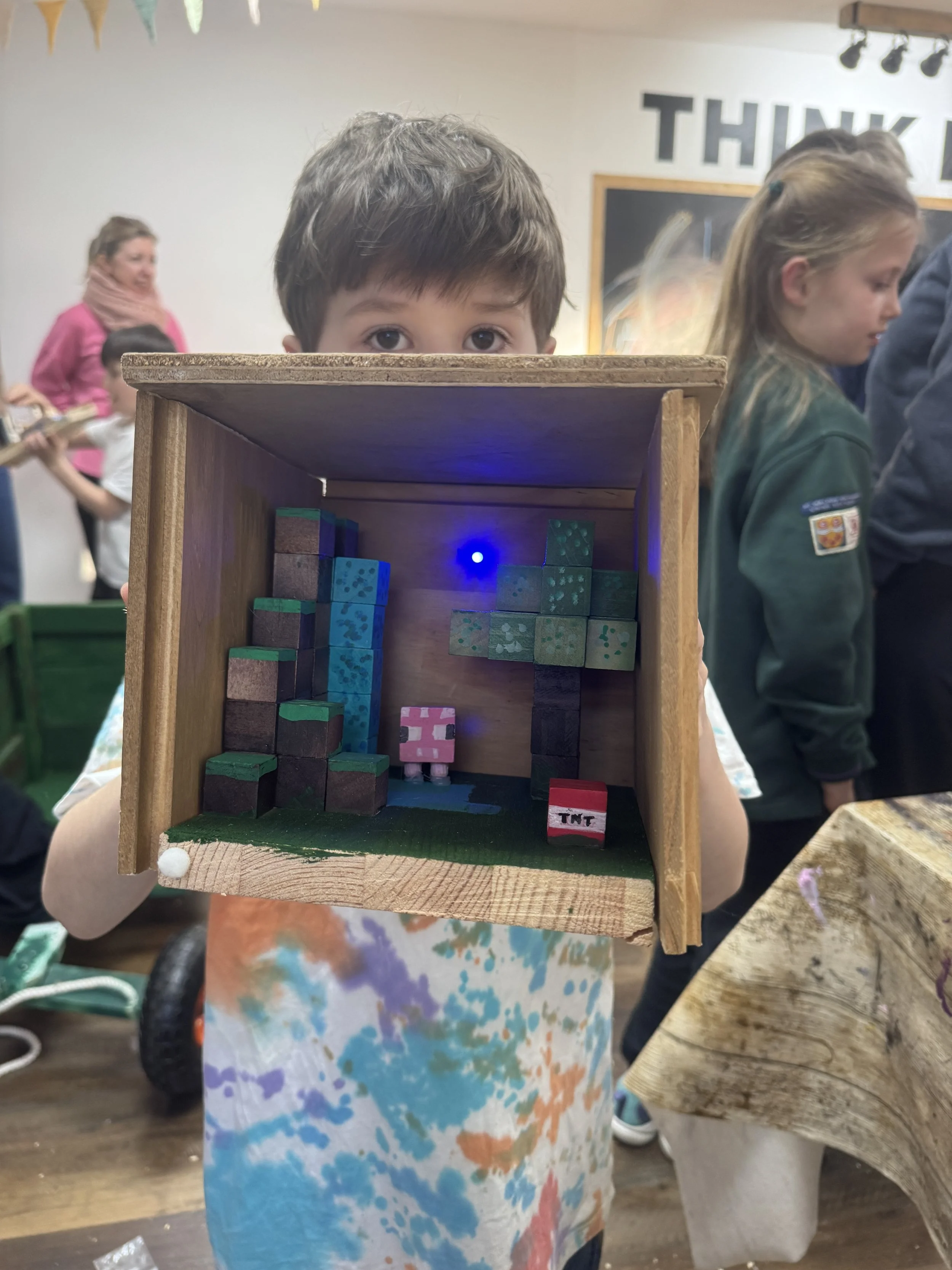 Child holding a wooden diorama with toy blocks, including a pixelated pig, TMT block, and pixel art tree, with a blue LED light inside, at an indoor event.