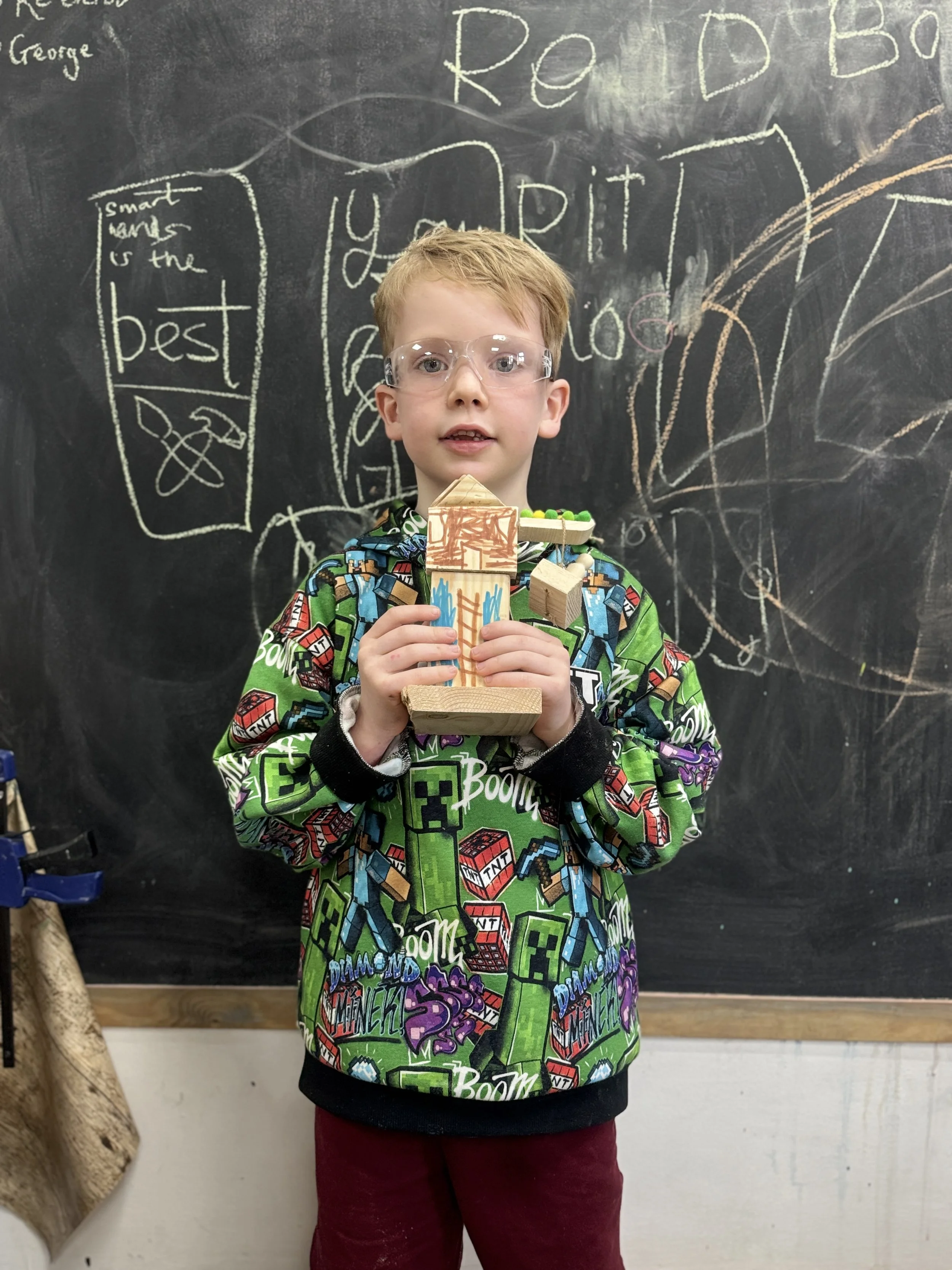A young boy wearing glasses and a colorful shirt with Minecraft characters stands in a classroom, holding a wooden craft model shaped like a building or tower, with a blackboard in the background covered in scribbles and writing.