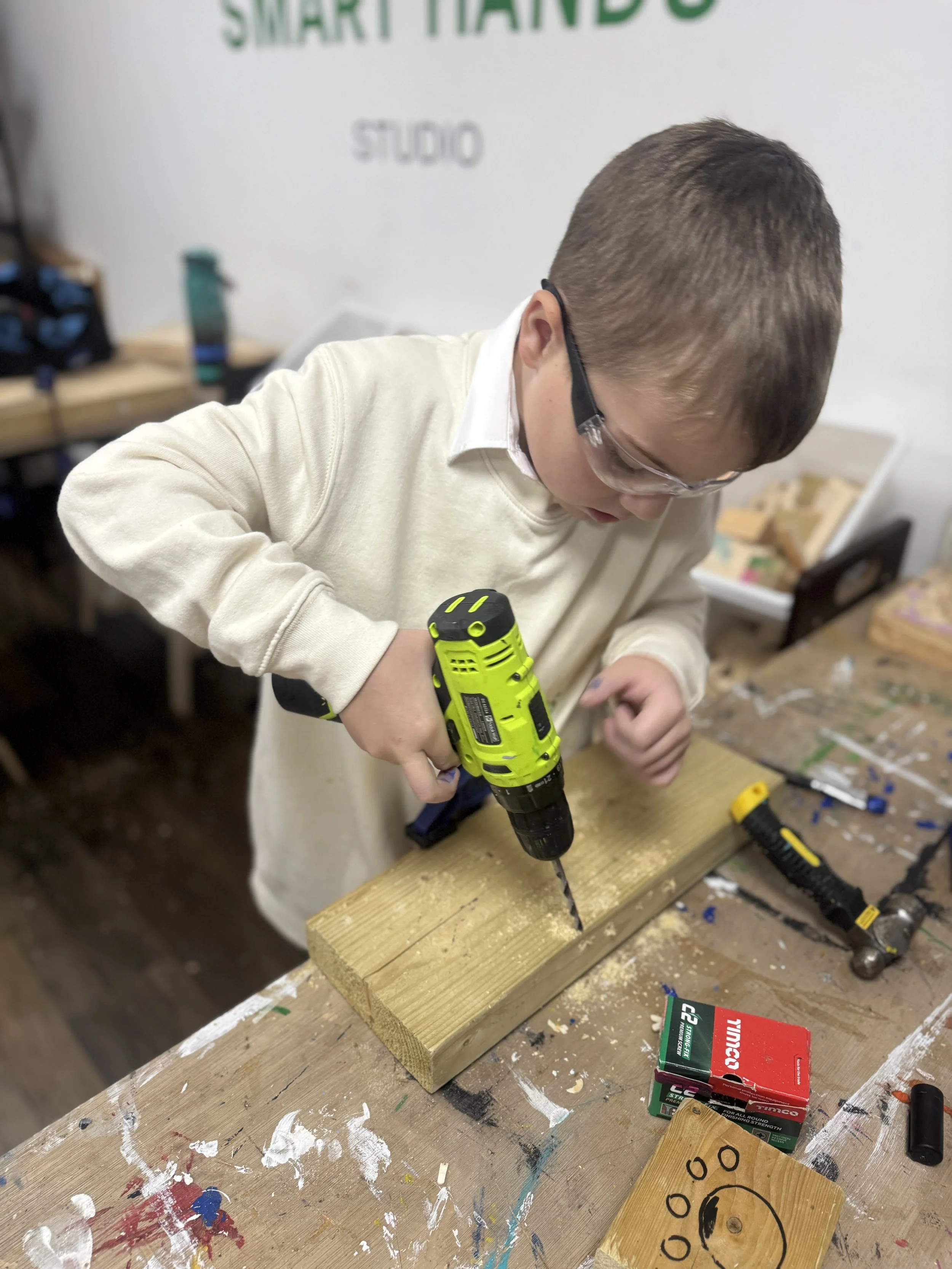 A young boy wearing glasses and a white sweater is drilling a hole into a piece of wood on a workbench, surrounded by tools and supplies.