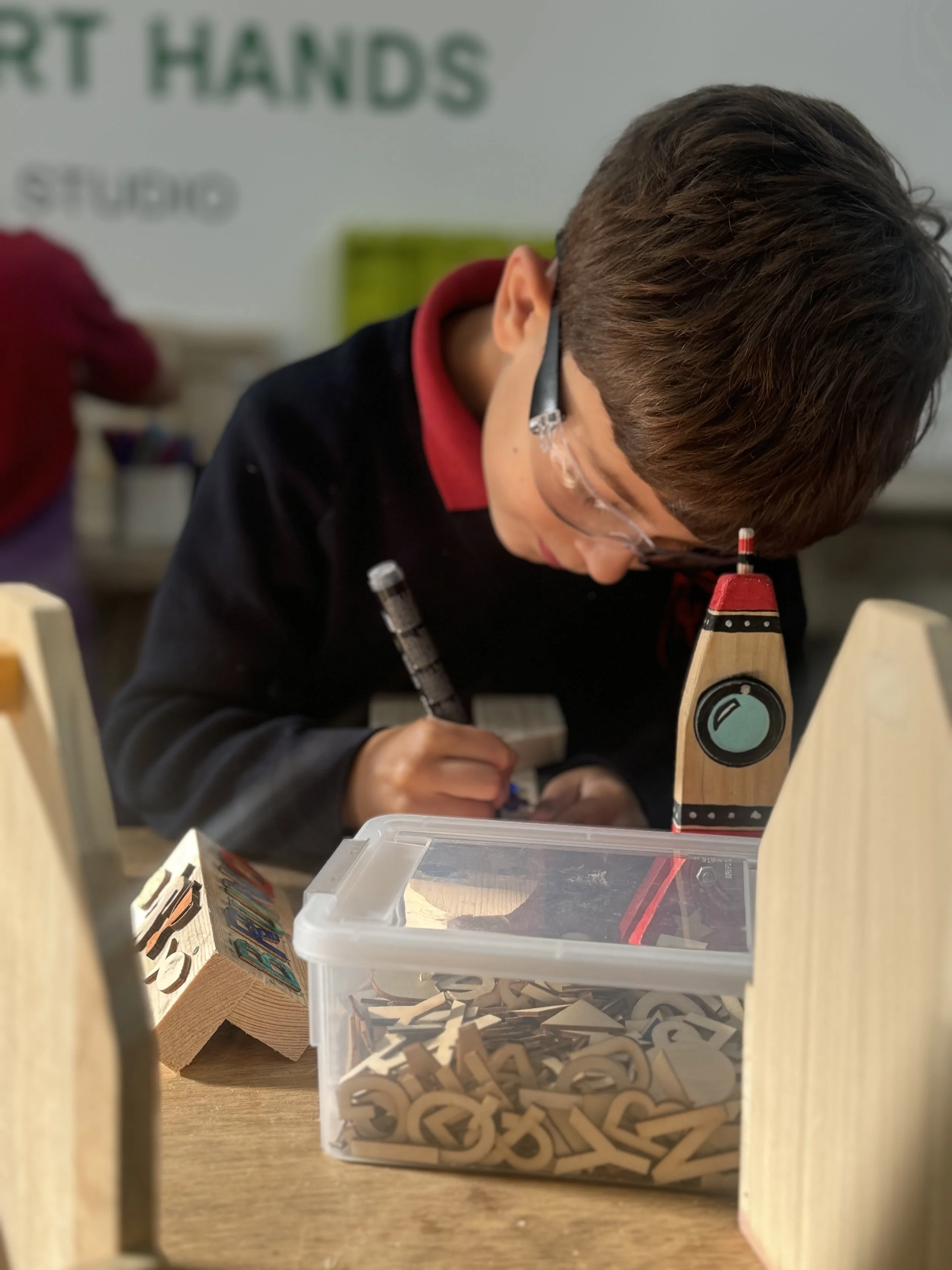 A young boy wearing safety glasses is working with wooden letters at a table, with a container of letter pieces in front of him and a painted wooden lighthouse nearby.
